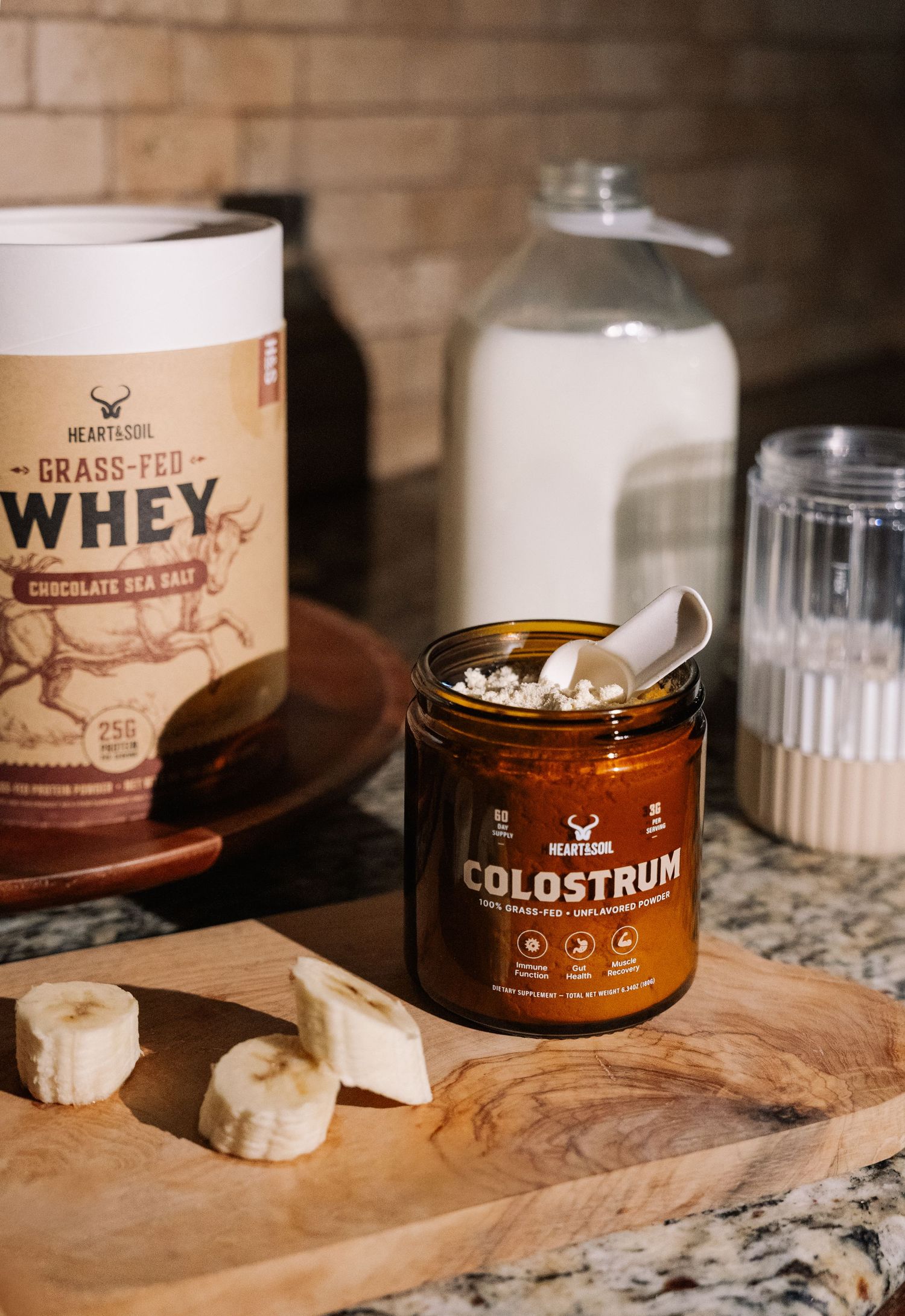Heart & Soil grass-fed whey protein chocolate sea salt and colostrum powder displayed on wooden cutting board with banana slices.