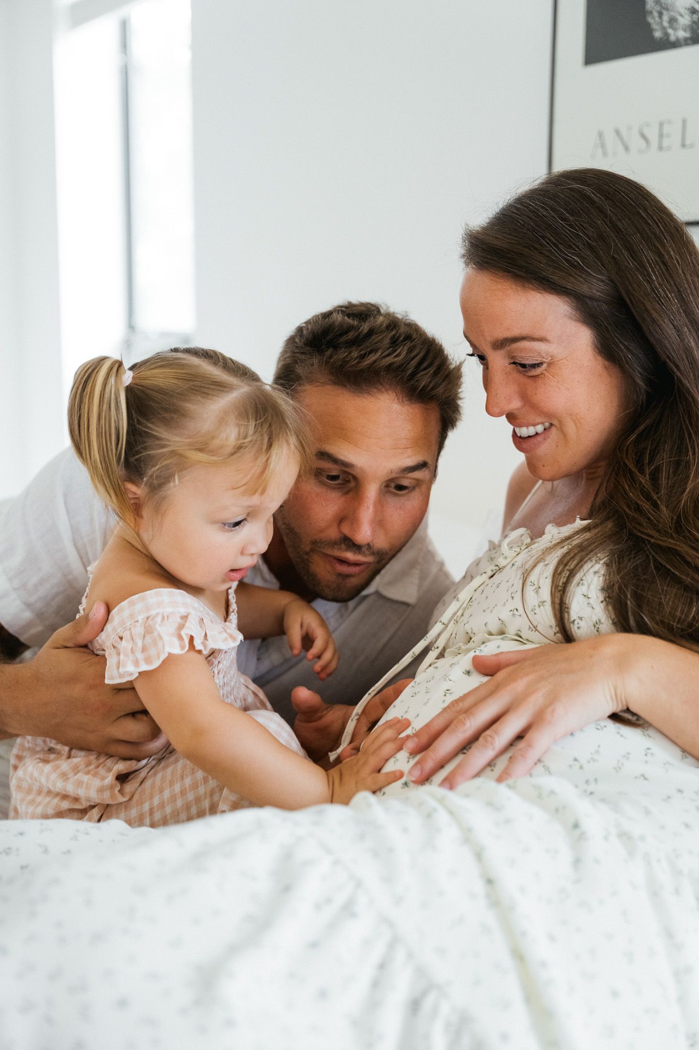 Pregnant woman smiling with husband and toddler daughter on white bed during maternity photo session.
