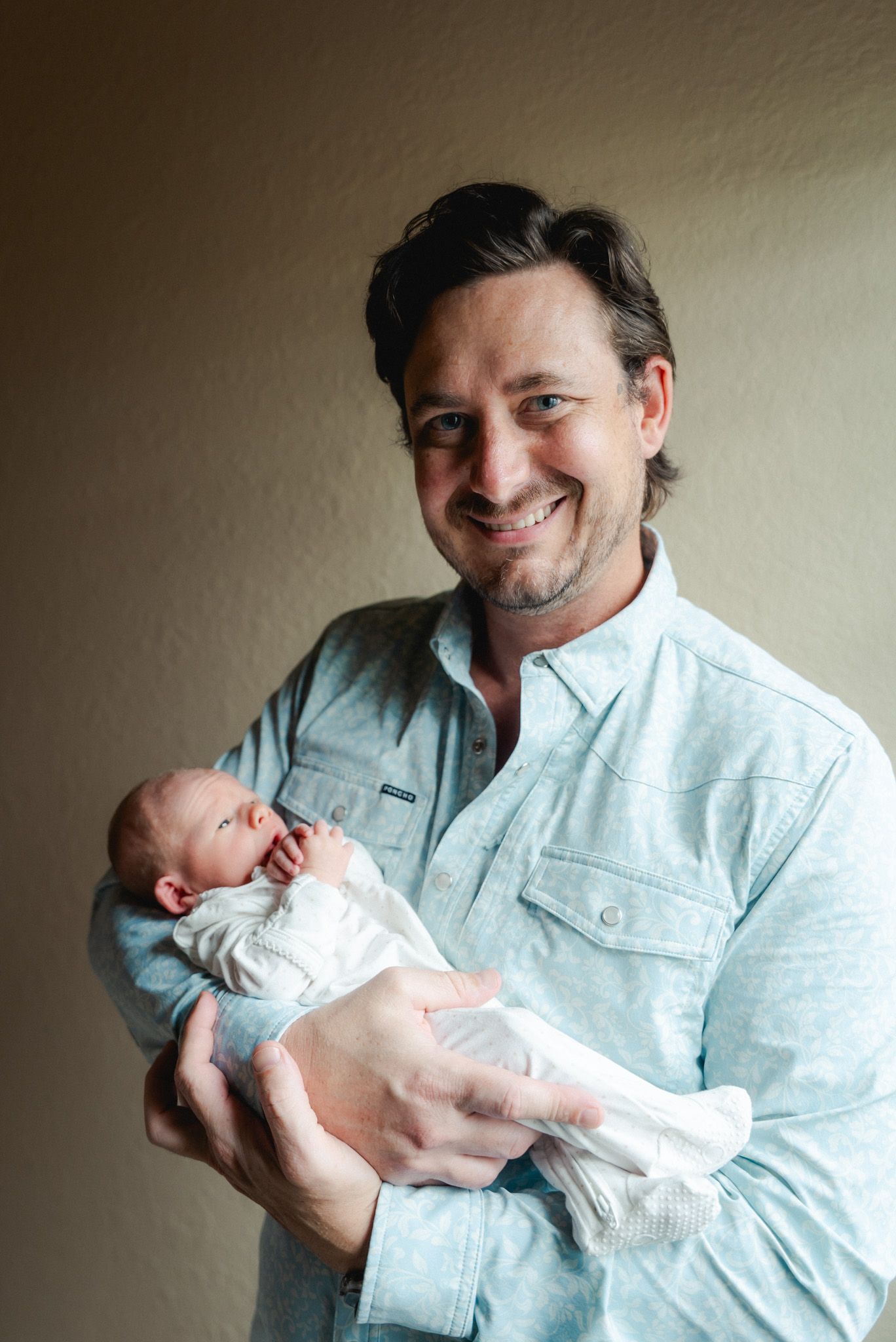 Smiling father in light blue shirt proudly holding swaddled newborn baby.
