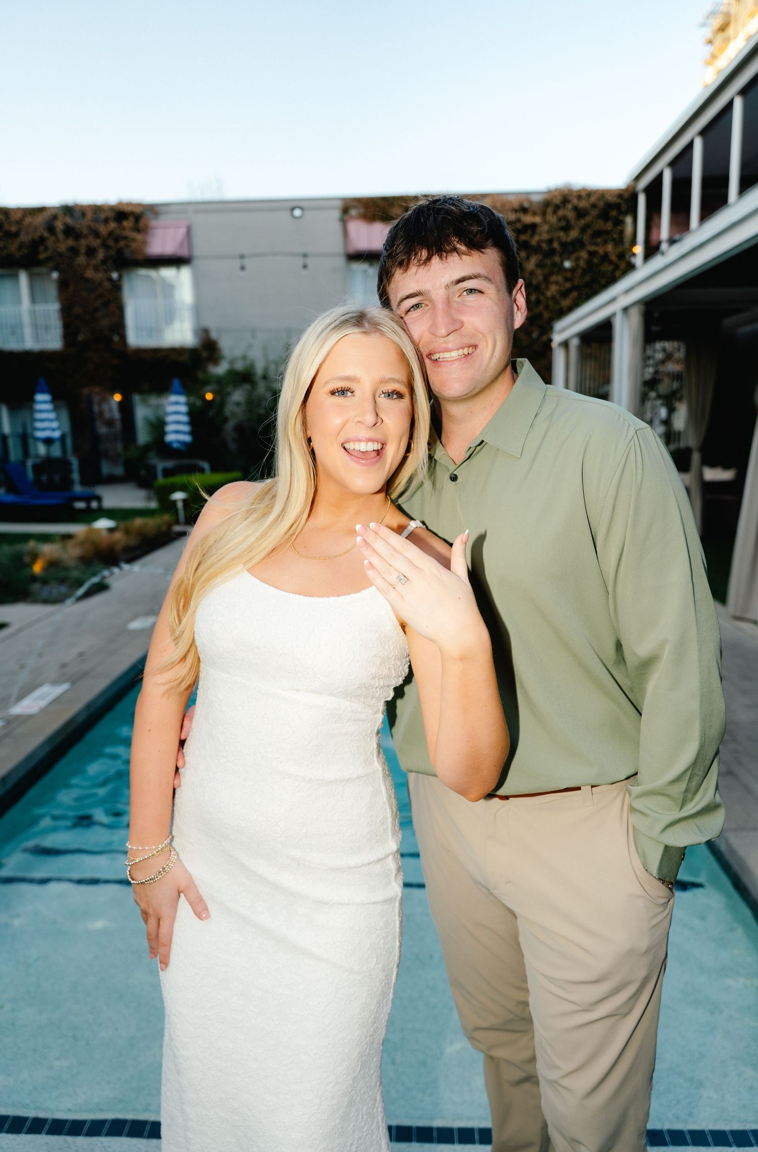 Engaged couple posing by pool, woman in white dress showing ring, man in sage green shirt, at outdoor venue.