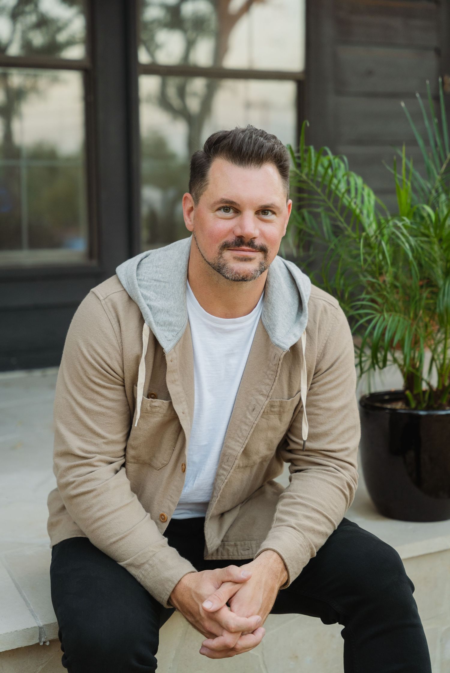 Smiling middle-aged man in beige hoodie jacket sitting outdoors near potted palm plant and modern building.
