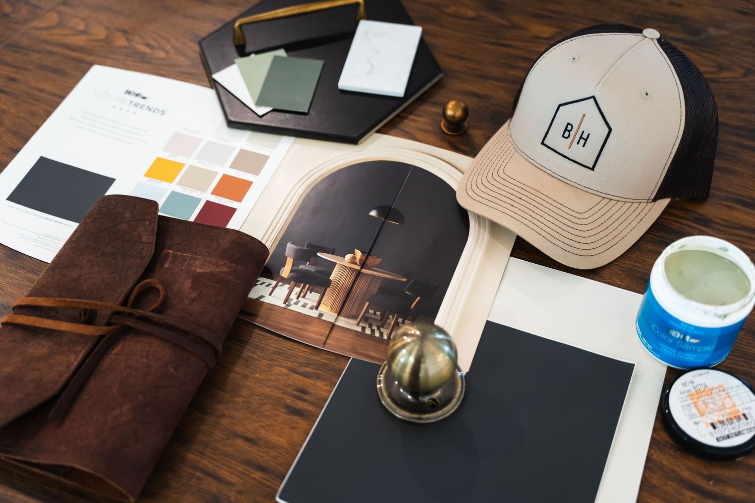 A flat lay of design materials including a color palette, cap, leather roll, tablet, and paint samples on wood.