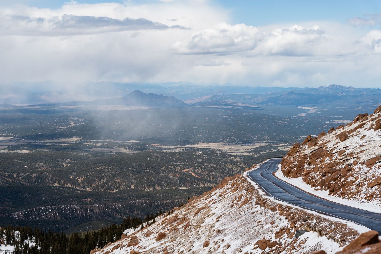 Aerial view of a winding mountain road with snow, overlooking a vast valley under dramatic cloudy skies.