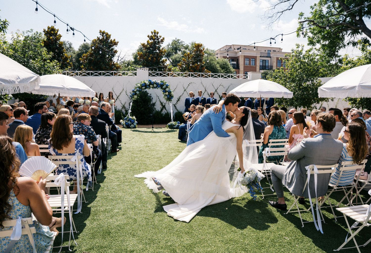 Couple shares first kiss as newlyweds during outdoor wedding ceremony on green lawn with white chairs and guests.