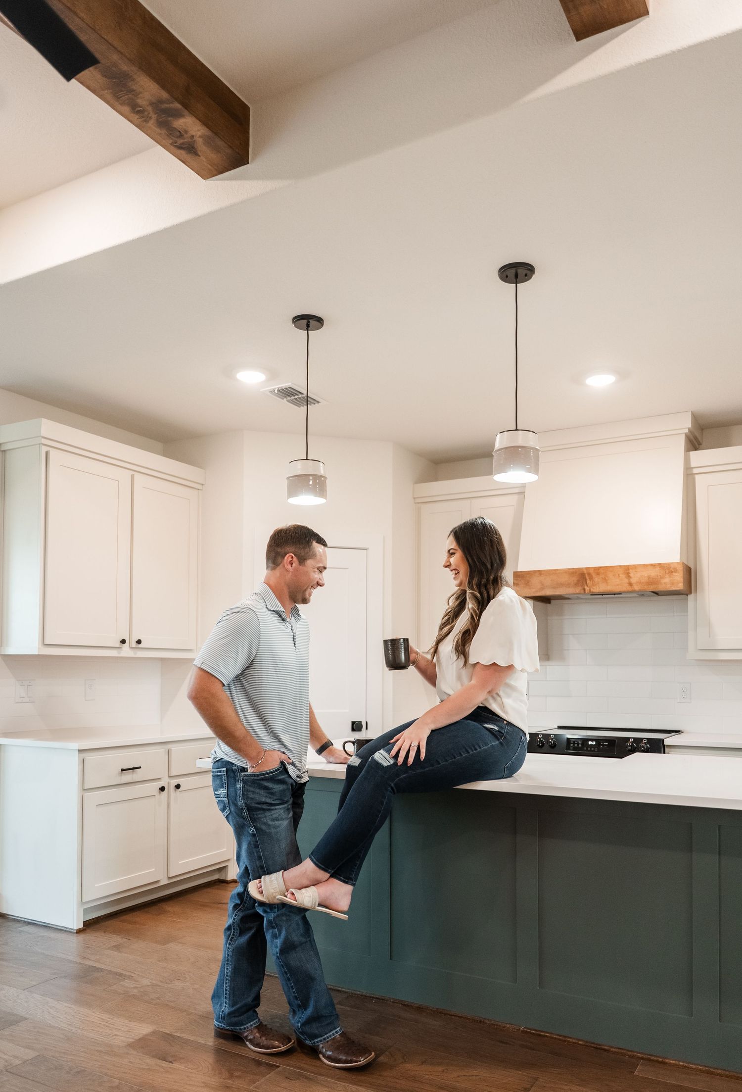 Couple chatting in a modern farmhouse kitchen with green island, white cabinets, and wood beam ceiling.