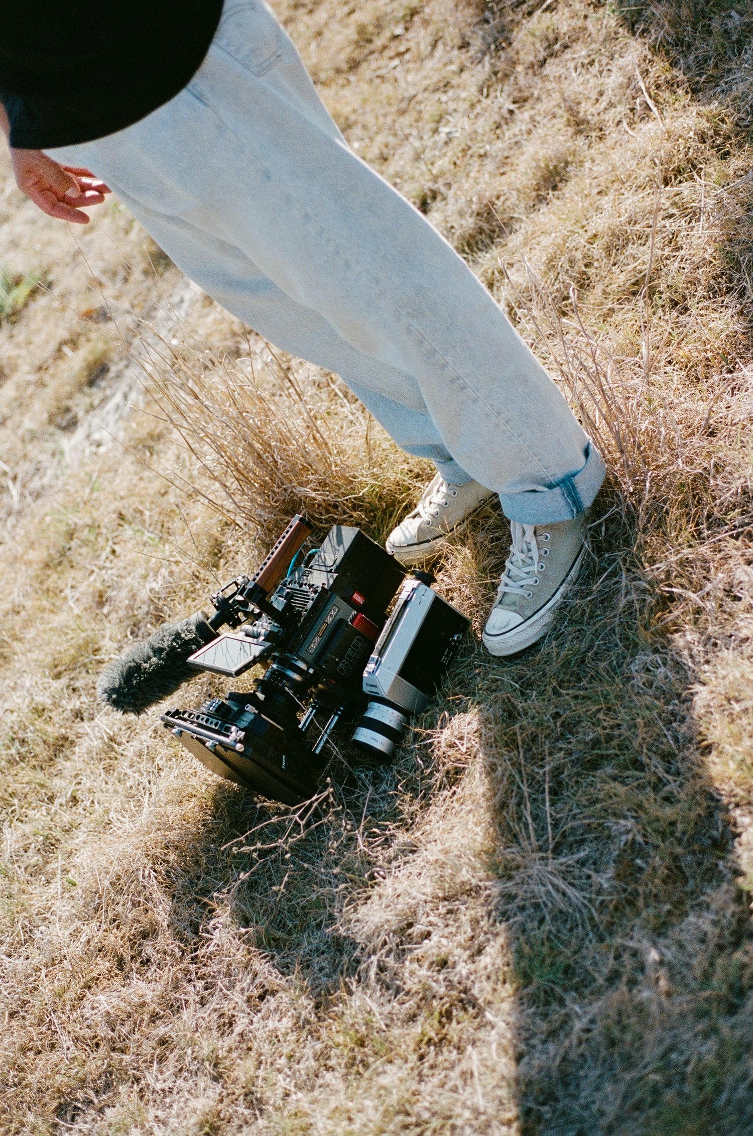 Professional video camera on dry grass beside person wearing white jeans and sneakers.