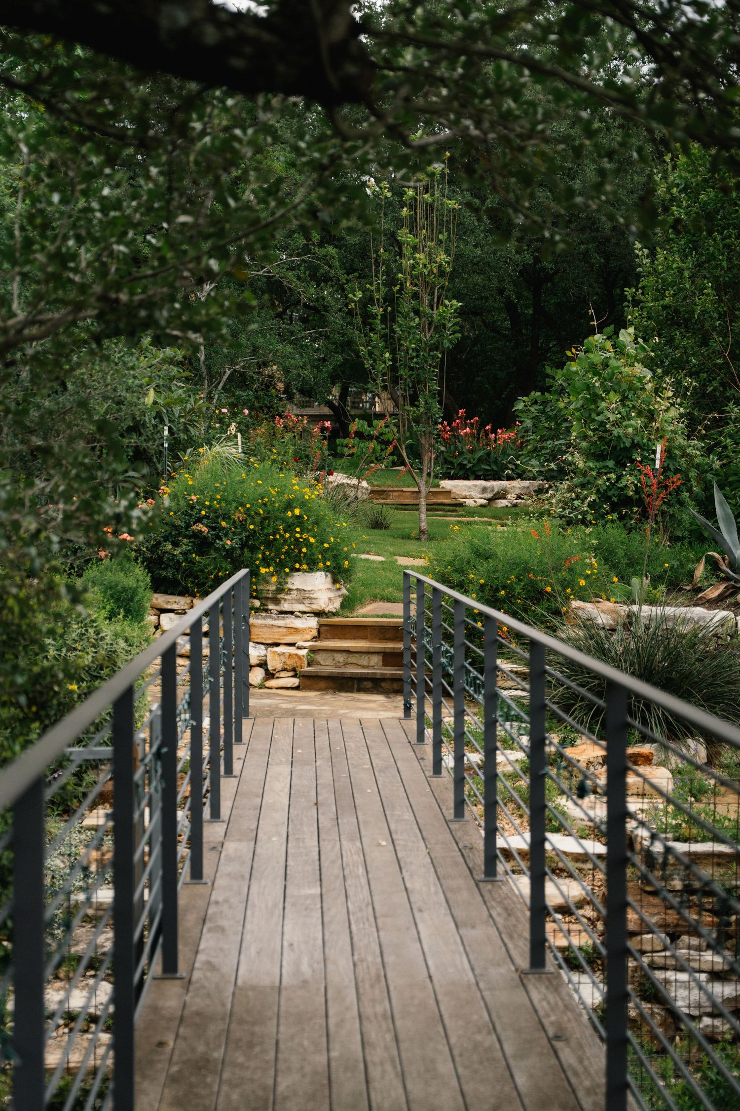 Wooden footbridge with metal railings leading to lush terraced garden with colorful wildflowers and mature trees.
