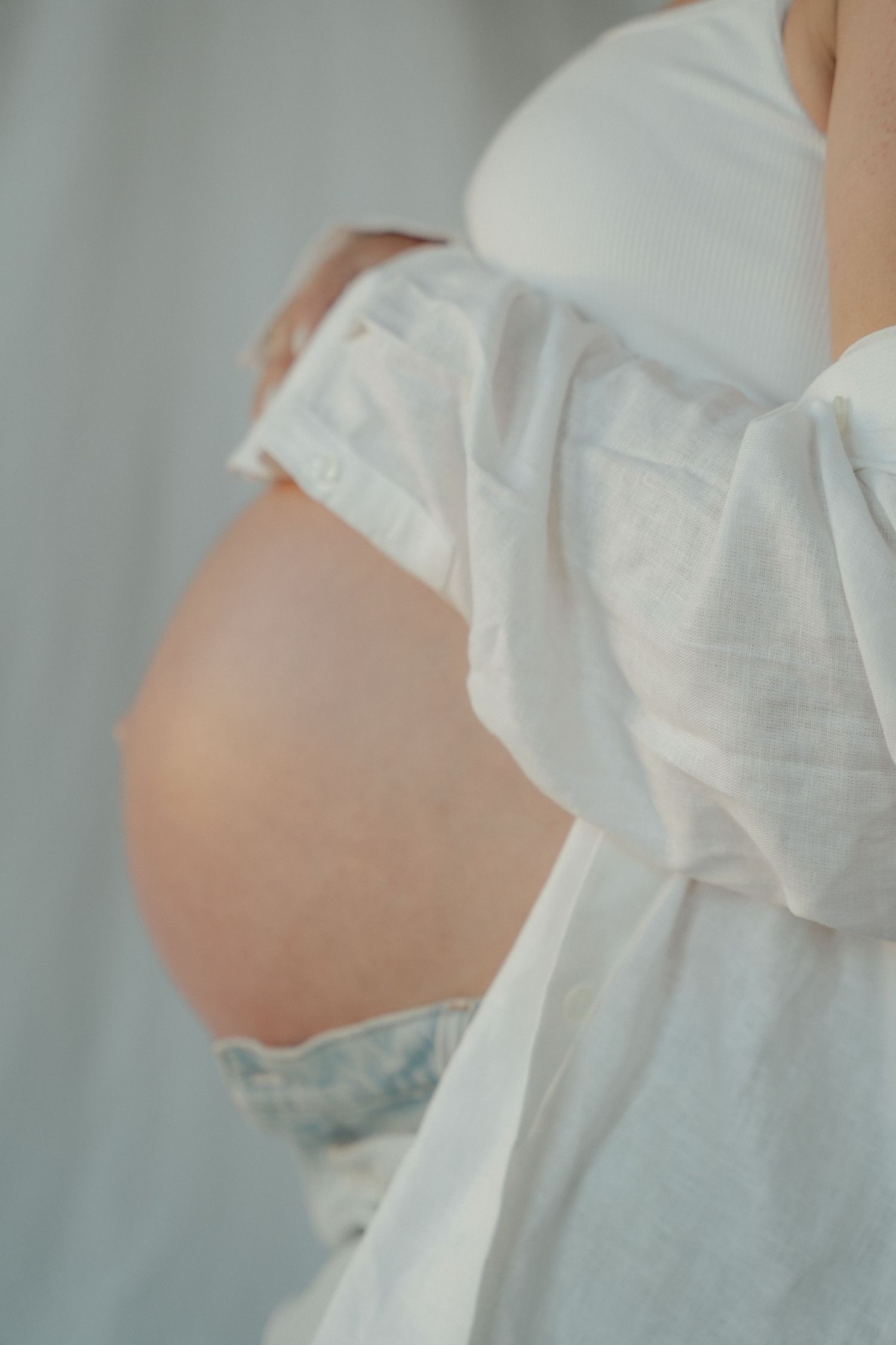 Close-up of a pregnant woman cradling her bare baby bump in a soft white dress.