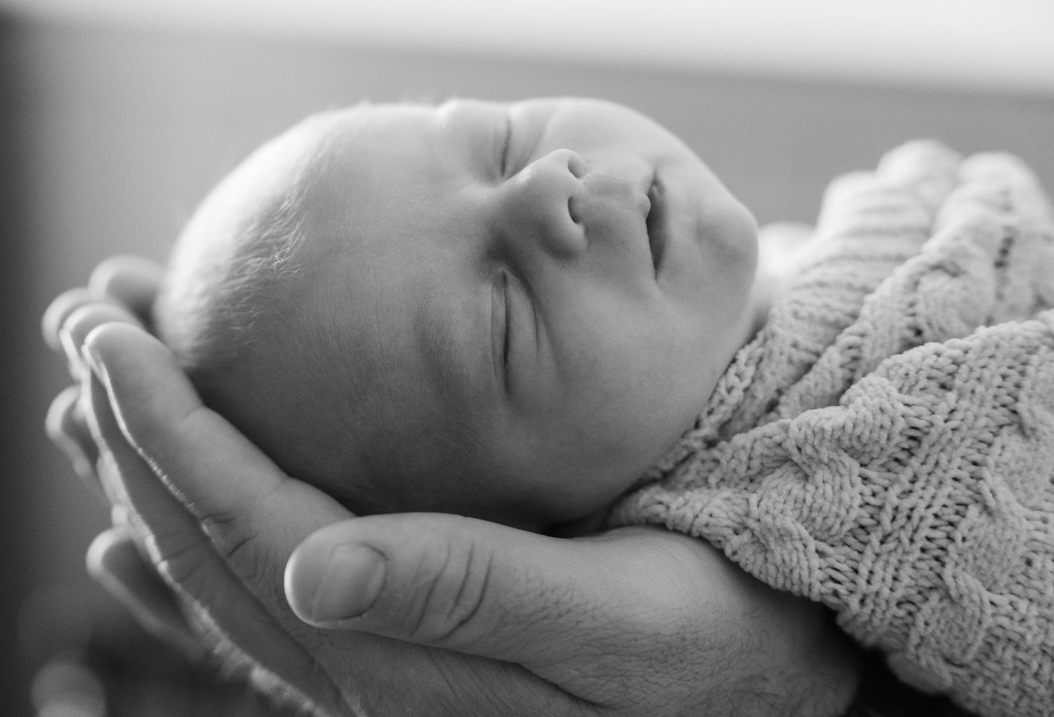 Newborn baby sleeping peacefully in adult hands wrapped in knit blanket, black and white photo.
