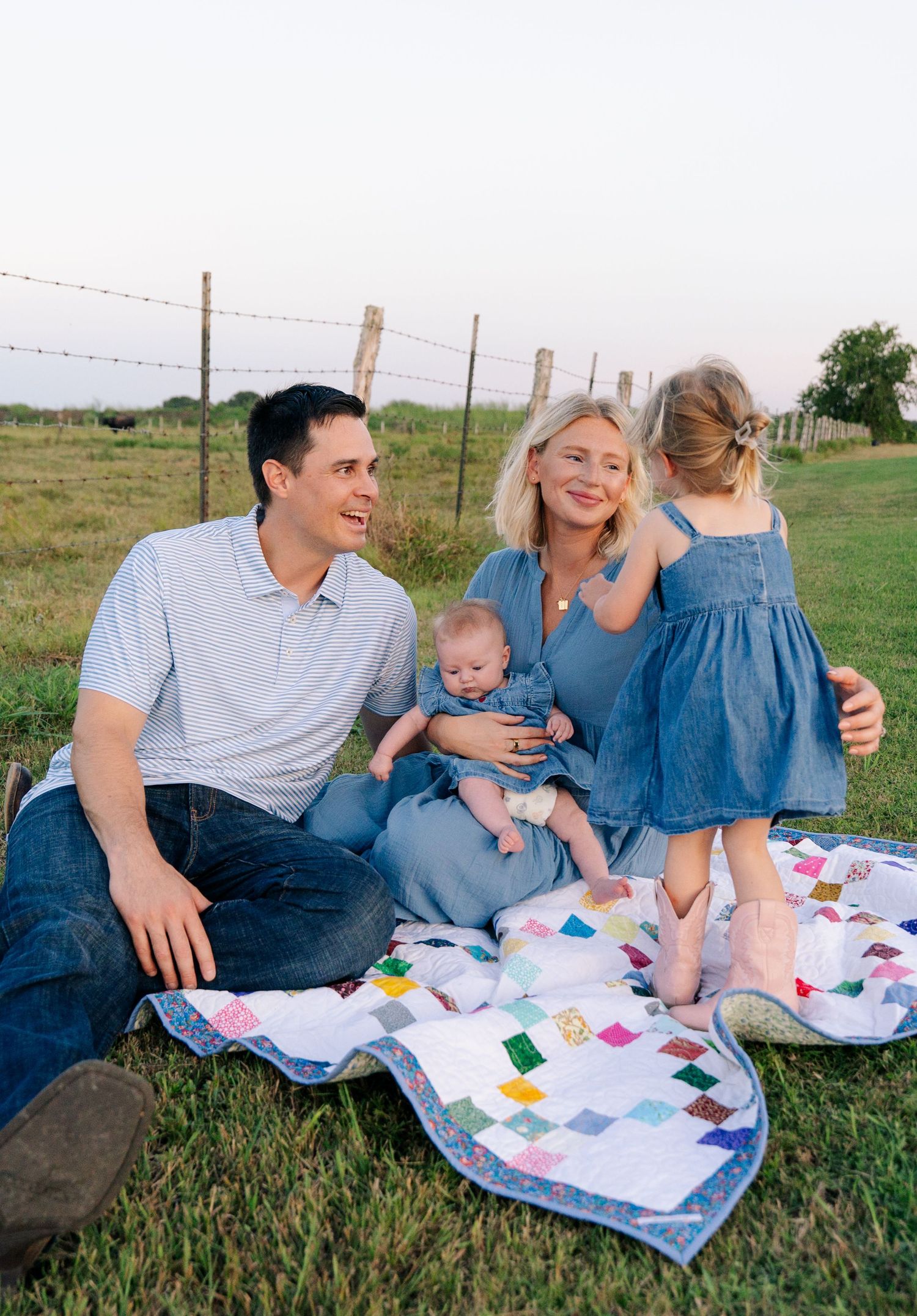 Happy young family with two children sitting on a colorful quilt in a green field at sunset.