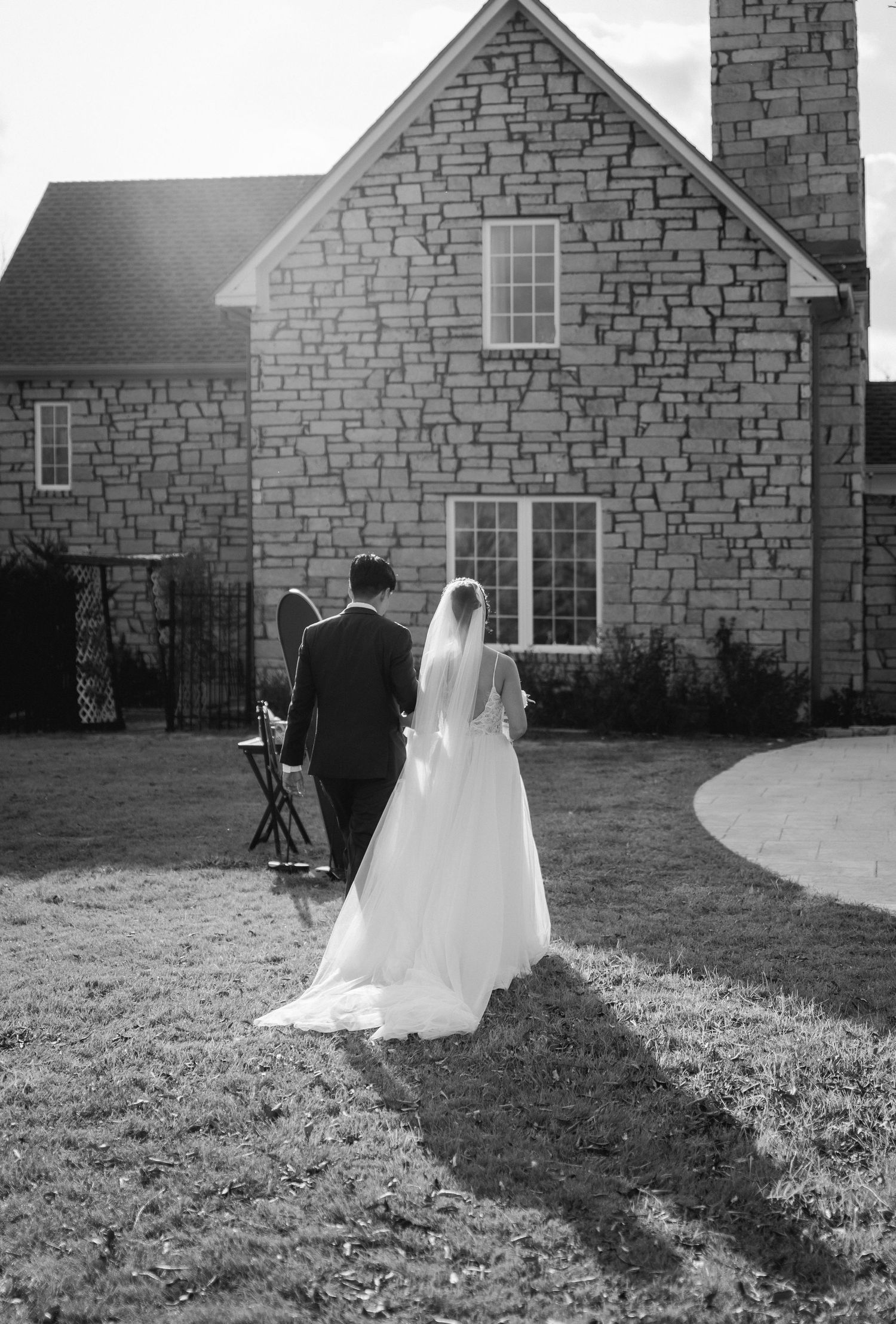 Black and white photo of bride and groom walking toward stone building on wedding day.