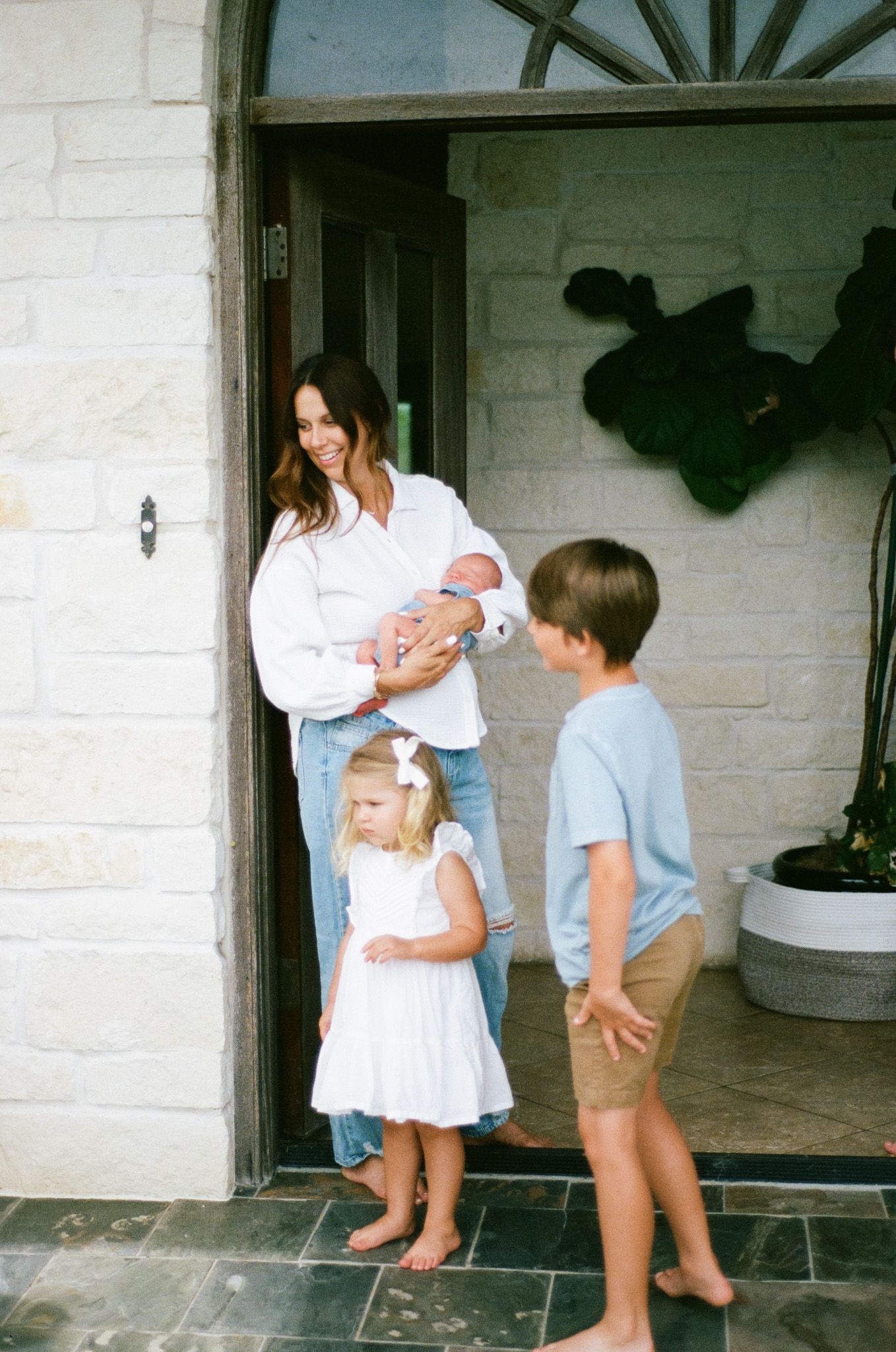 A smiling brunette mother holds her newborn in a doorway as her young daughter and son look on with curiosity.