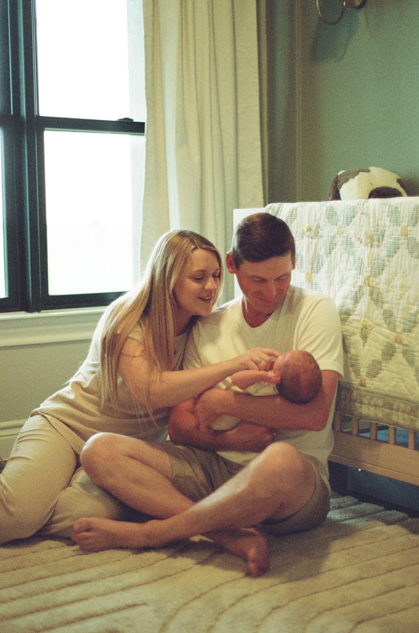 Happy new parents sitting on nursery floor together admiring their precious newborn baby by crib.