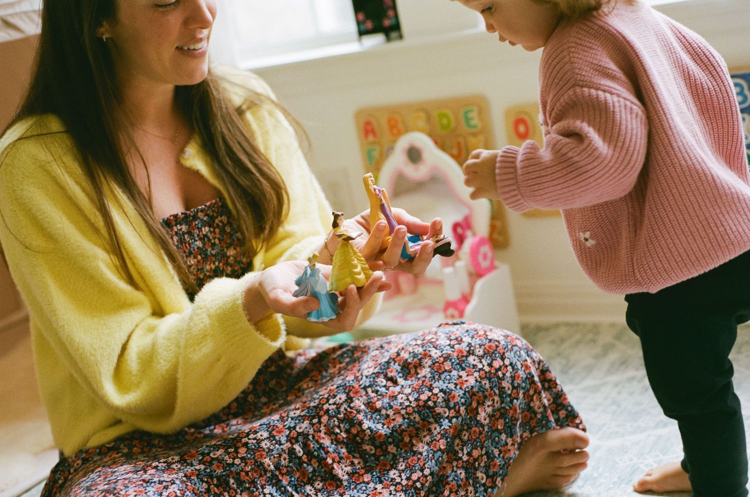 A mother in a yellow cardigan sits on the floor playing with colorful toys with her young toddler.
