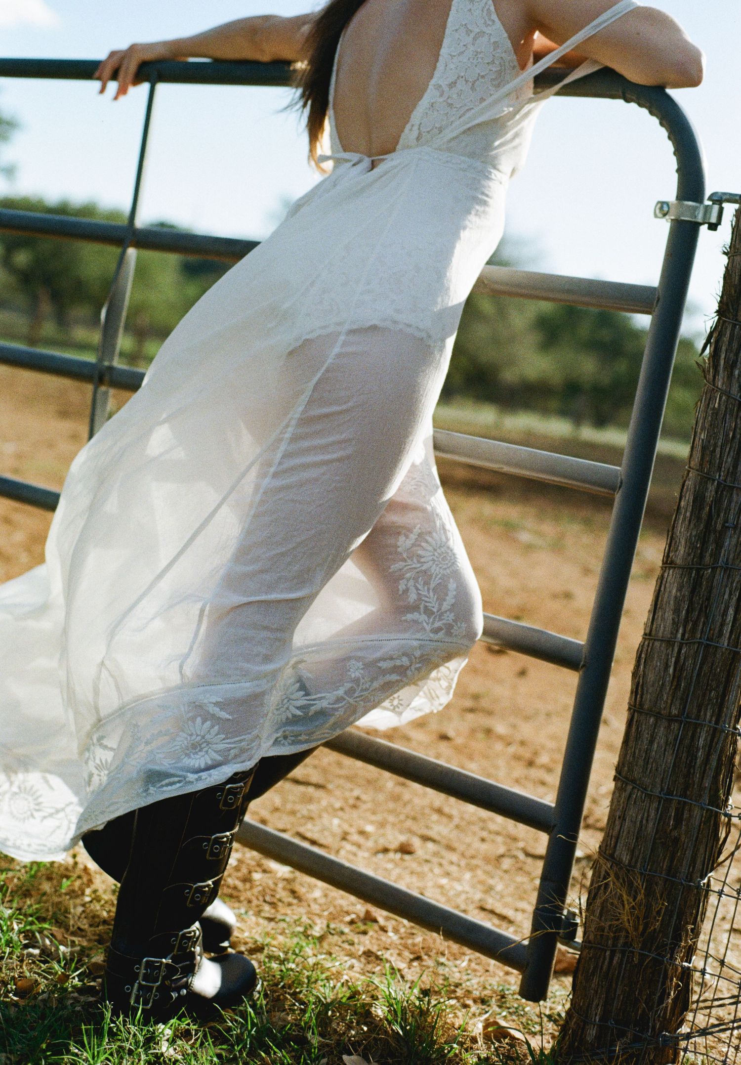Woman in white lace dress and cowboy boots climbing a metal farm gate outdoors.