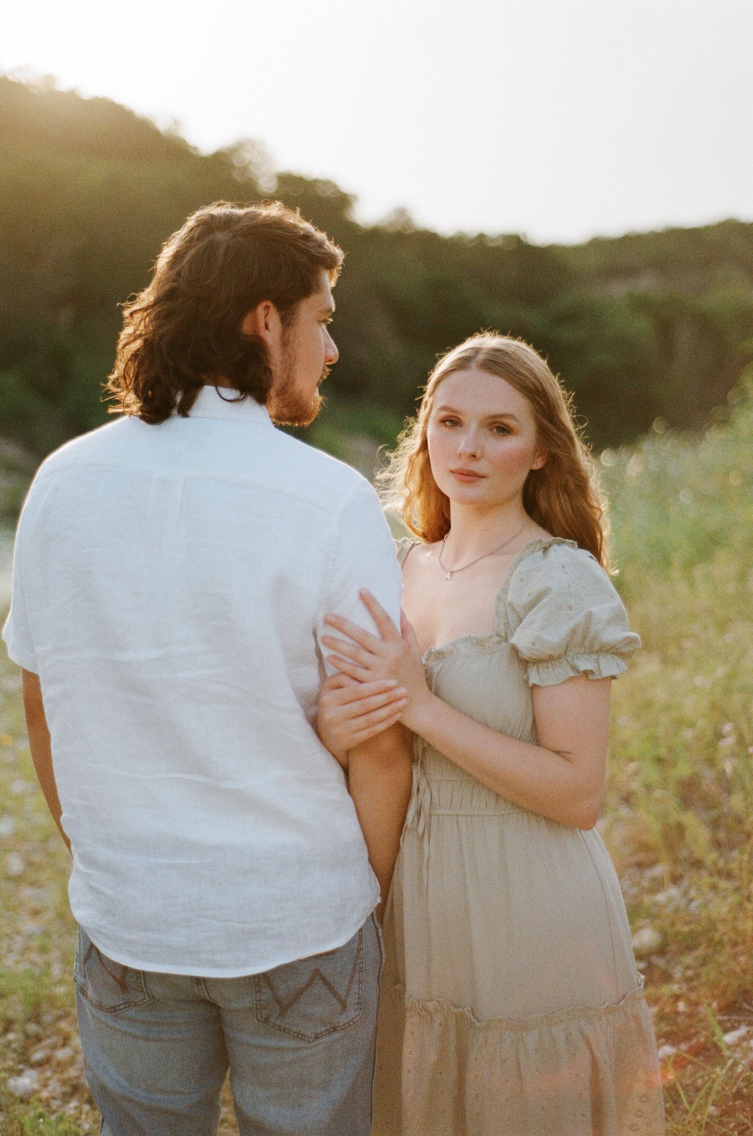 A couple shares a tender moment in a sunlit field, the woman in a sage dress gazing forward as golden hour glows.