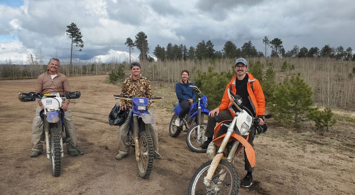 Four dirt bike riders posing with motorcycles on a sandy trail surrounded by pine trees and cloudy sky.