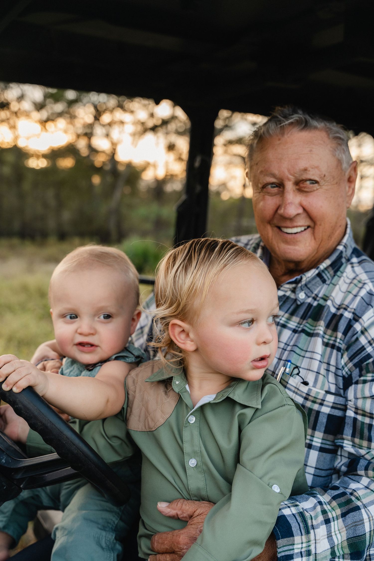 A cheerful grandfather smiles warmly while sitting with two young grandsons in a golf cart at sunset.