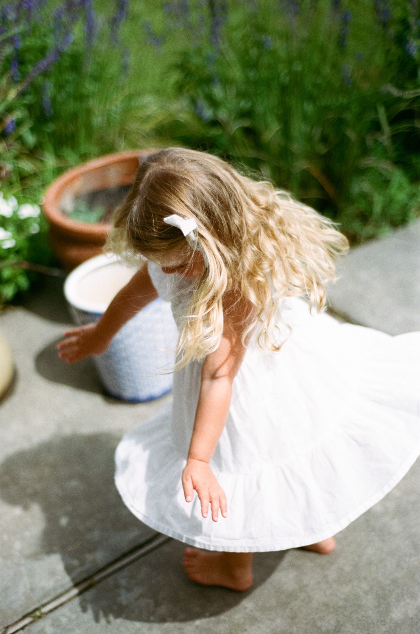 Young girl in white dress twirling barefoot in garden with potted plants and lavender.