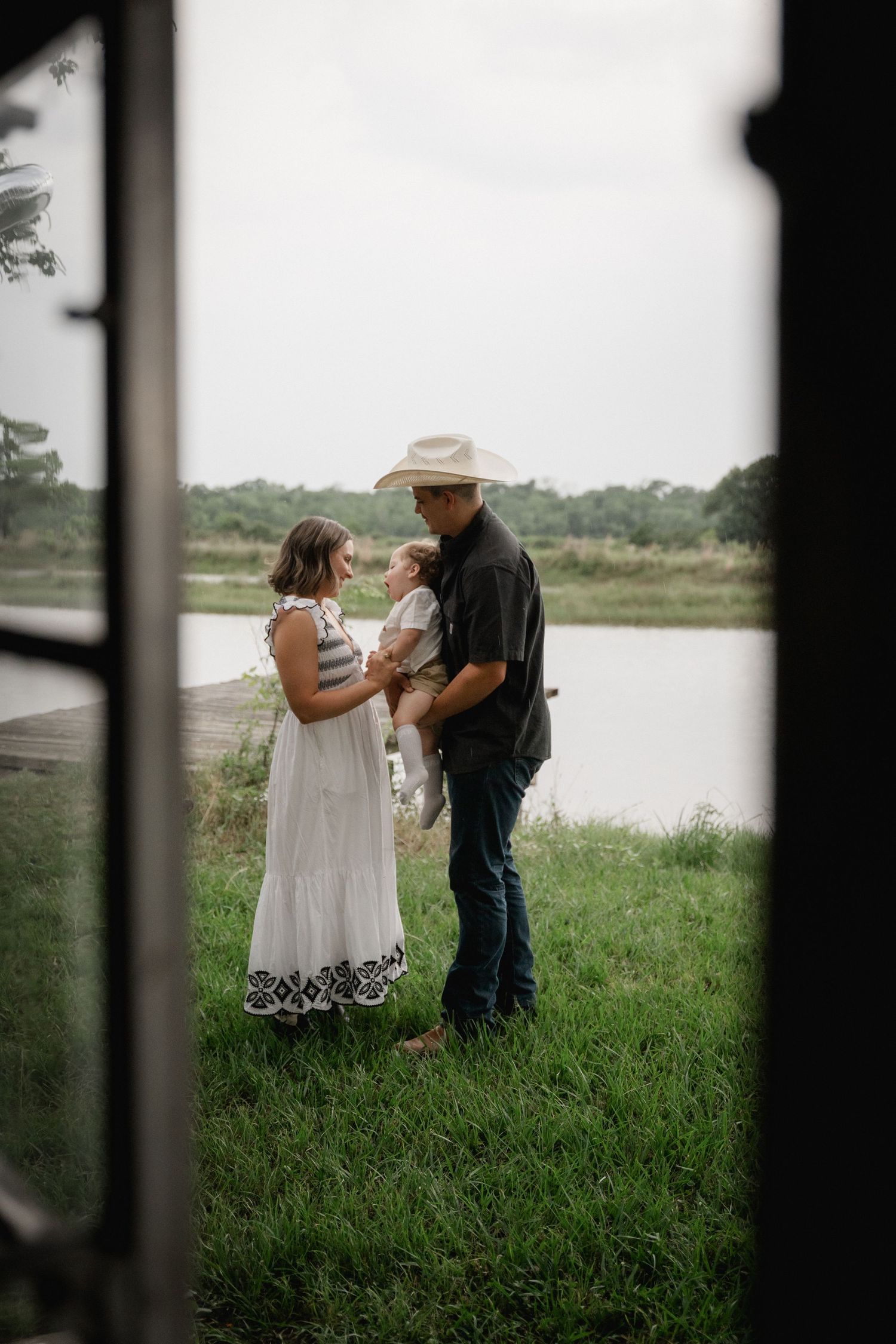 Family of three with baby standing by a tranquil lake, viewed through an open doorway.