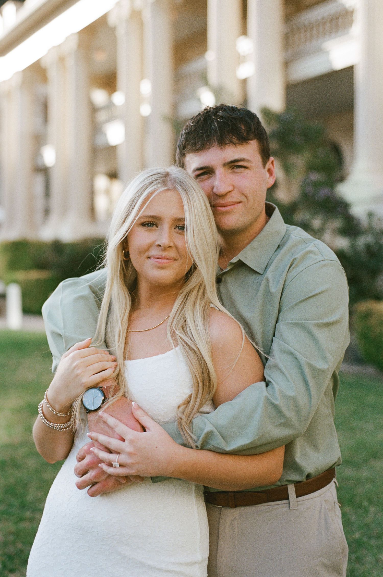 Man in sage shirt embracing woman in white dress from behind, showing engagement ring, near columns.