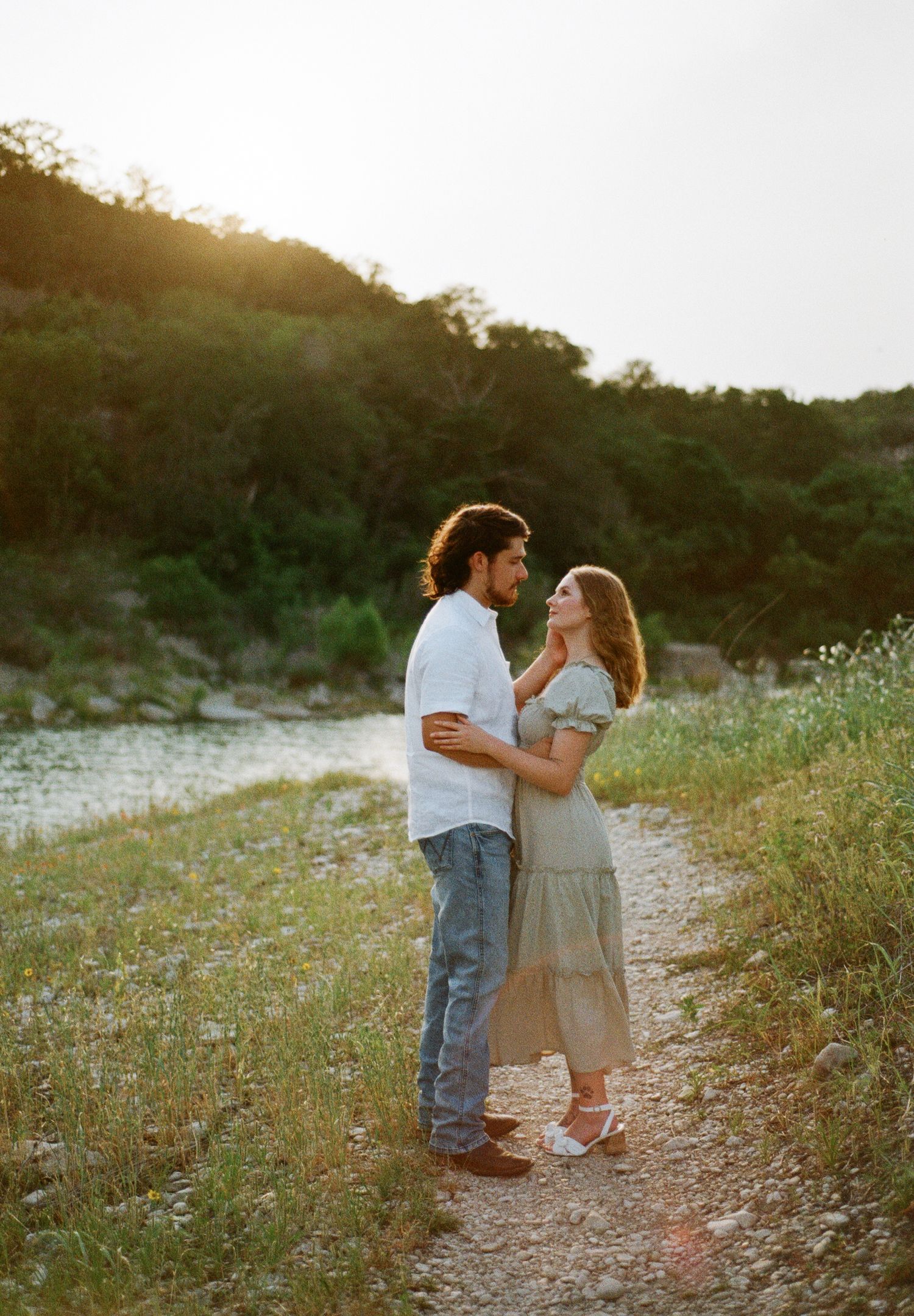 Couple sharing a romantic moment on a sunlit path near a river surrounded by lush greenery.