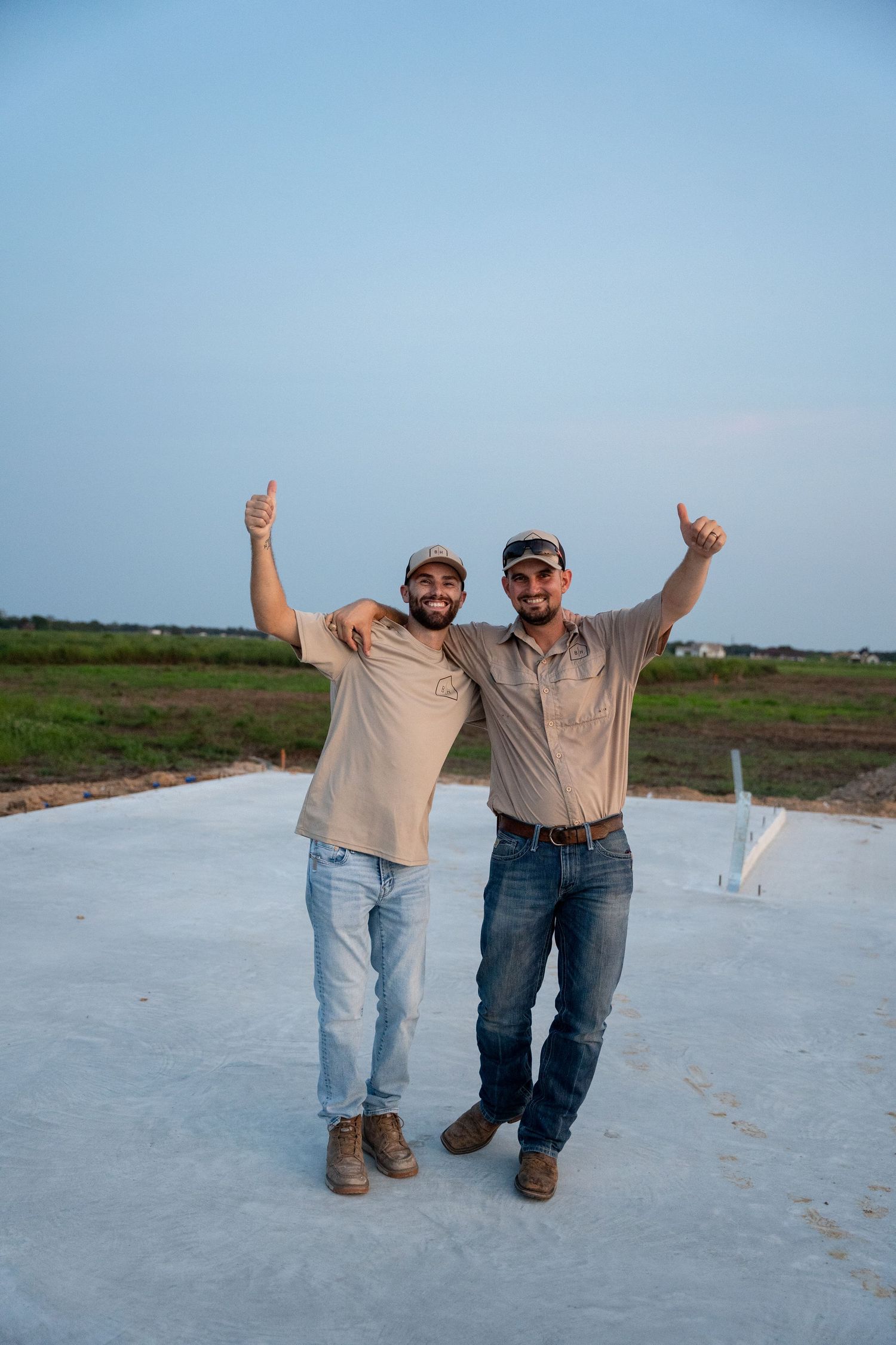 Two men in caps give thumbs up on a freshly poured concrete slab at a rural construction site.