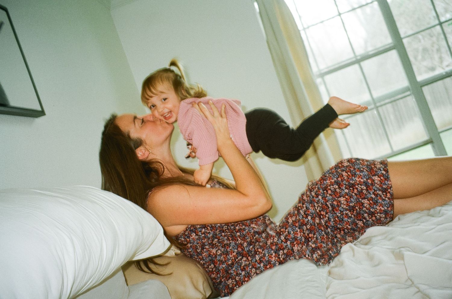 A joyful mother lies on a bed lifting her laughing toddler daughter playfully into the air.