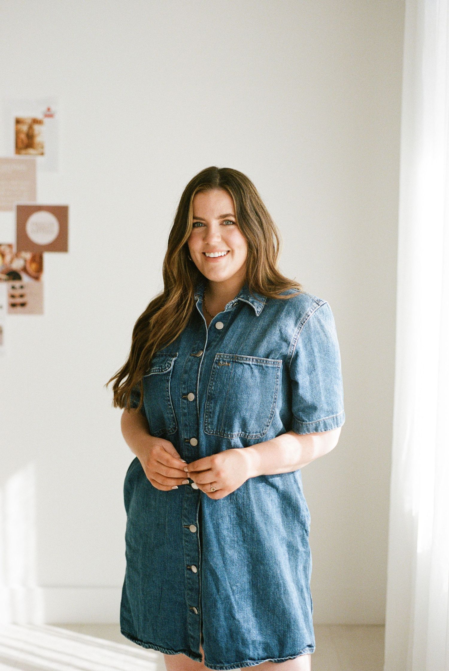 Smiling woman in a denim shirt dress standing in a bright white room.