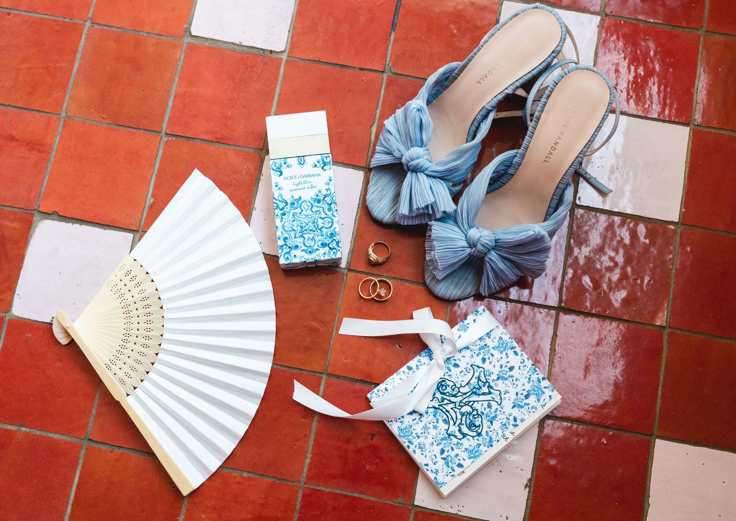 Flat lay of blue bow heels, white hand fan, gold rings, and blue floral accessories on terracotta tile floor.