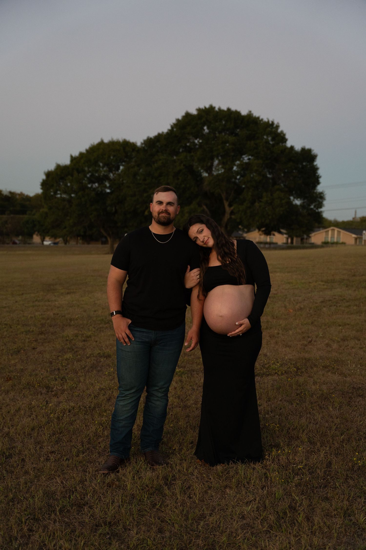 Couple maternity photo in field at golden hour, woman in black gown leaning on mans shoulder.