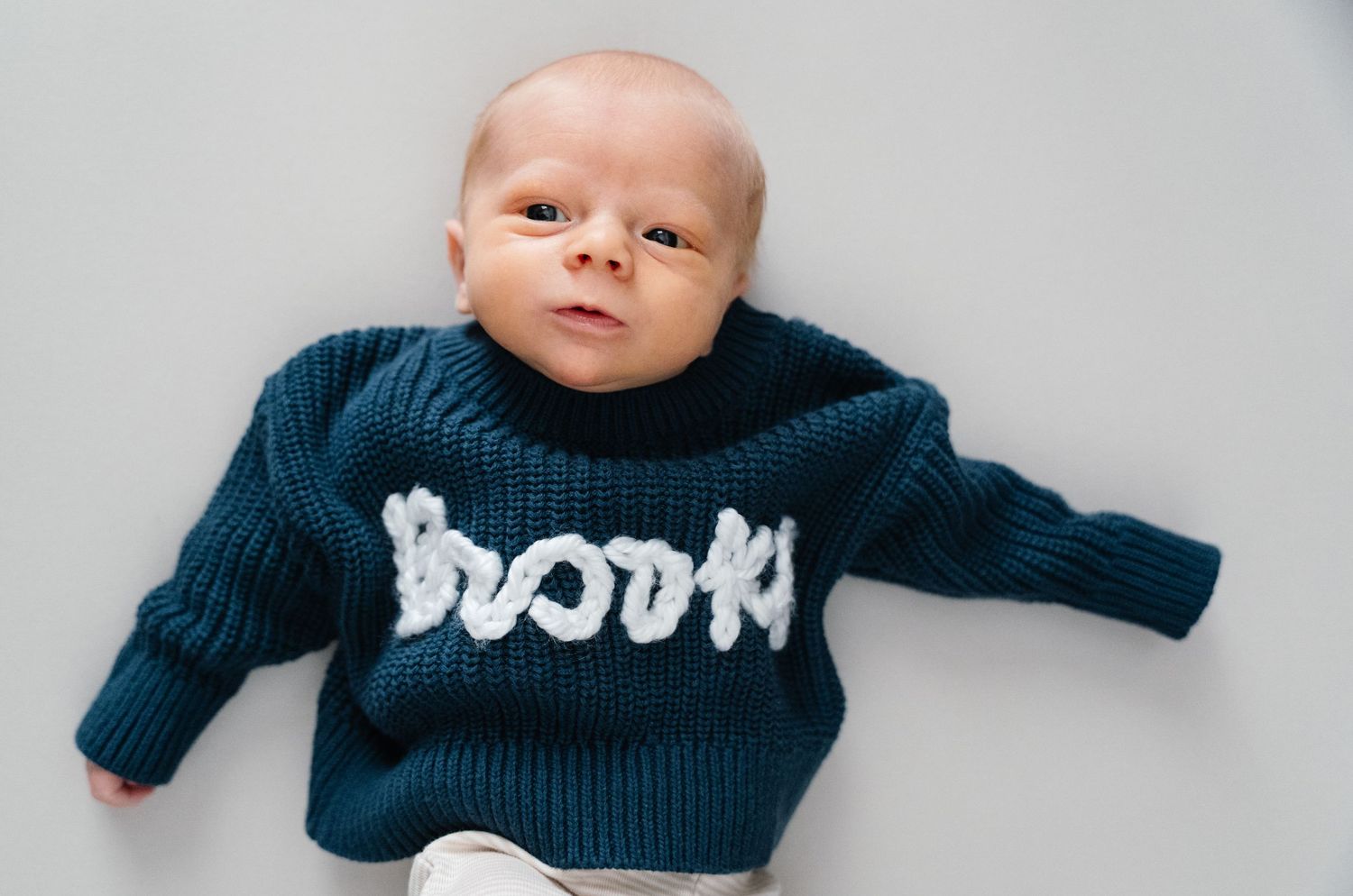 Close-up of newborn baby in teal knit personalized sweater lying on light gray background.