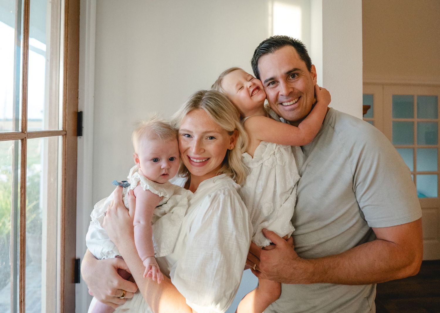 Smiling family of four posing indoors, parents holding two young daughters in a bright room.