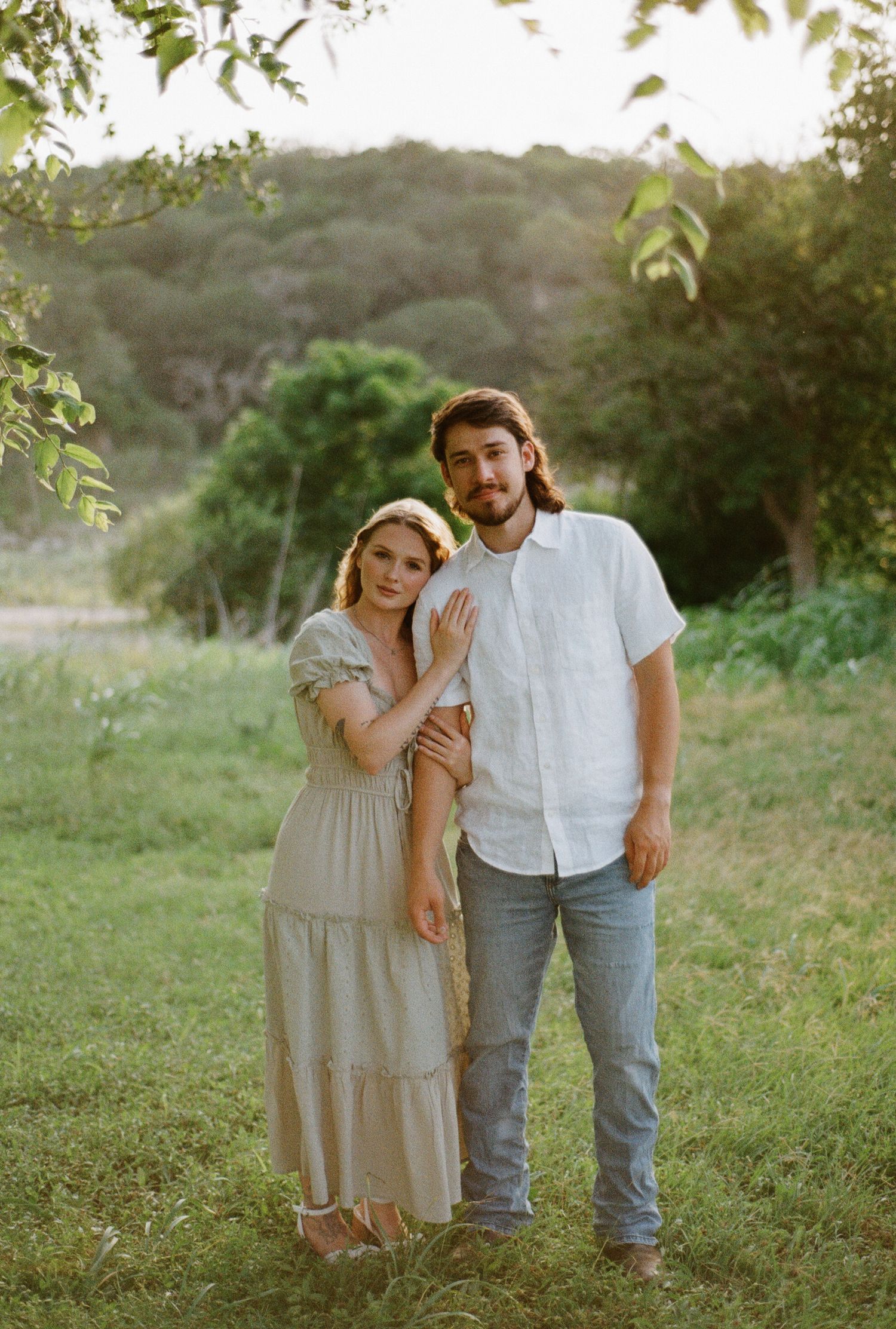 A couple poses in a lush green field, woman in beige dress leaning on man in white shirt and jeans.