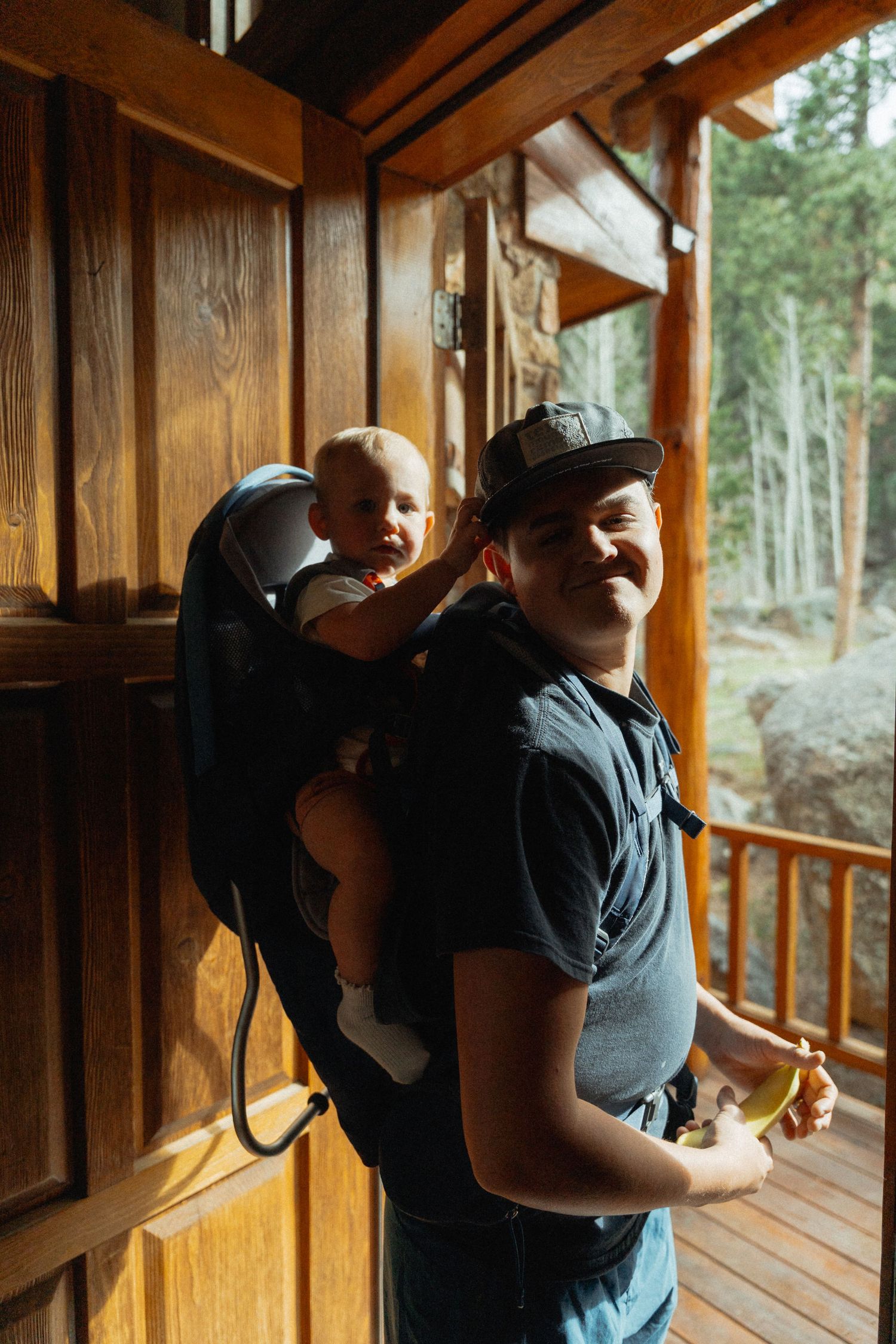 Smiling man wearing a cap carries a happy baby in a backpack carrier on a wooden cabin porch.