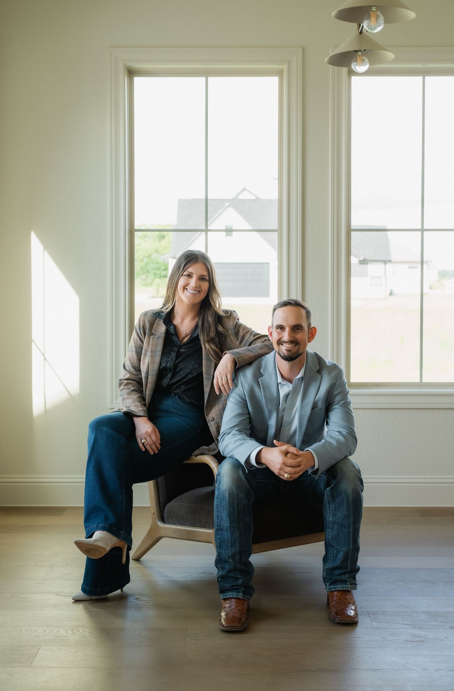 A man and woman pose together in a bright, modern room with large windows and hardwood floors.