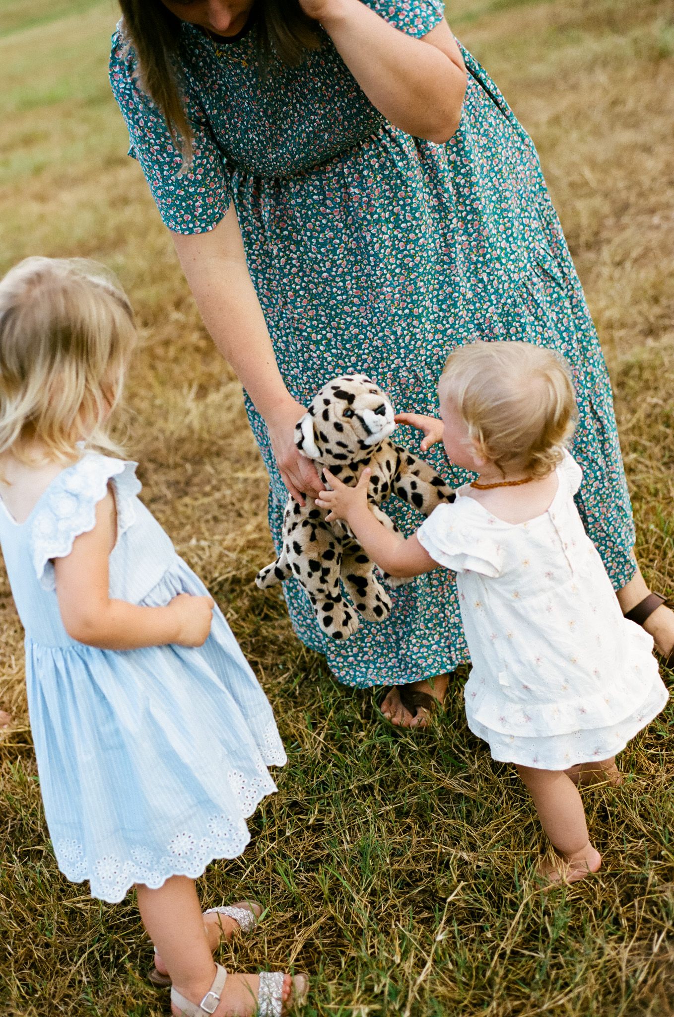 A mother in a floral dress hands a spotted stuffed animal to two young girls in white and blue dresses outdoors.