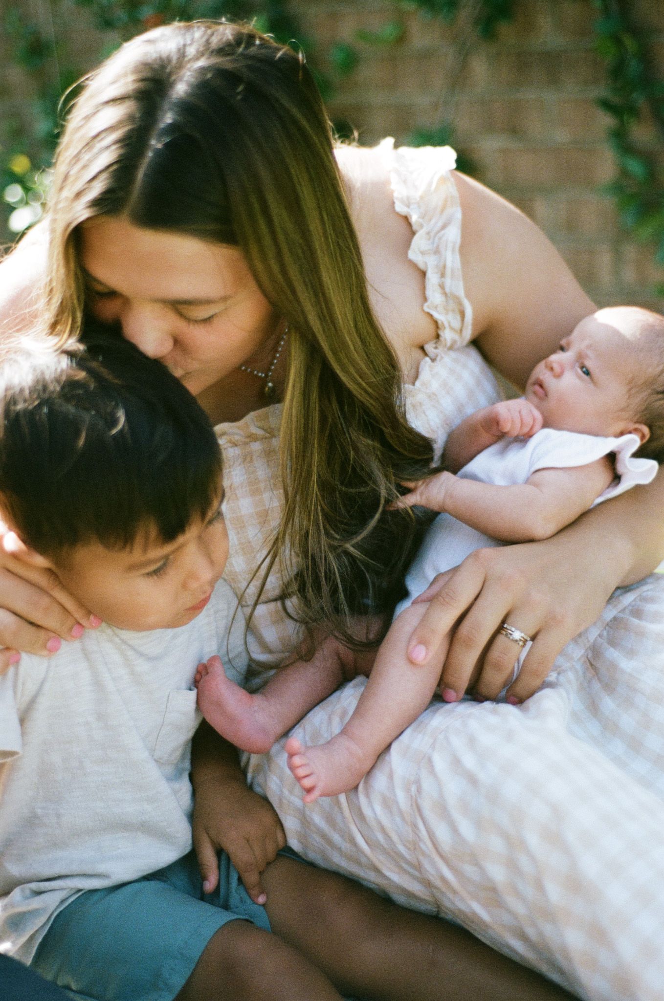 A mother tenderly kisses her toddler while cradling her newborn baby outdoors in warm sunlight.