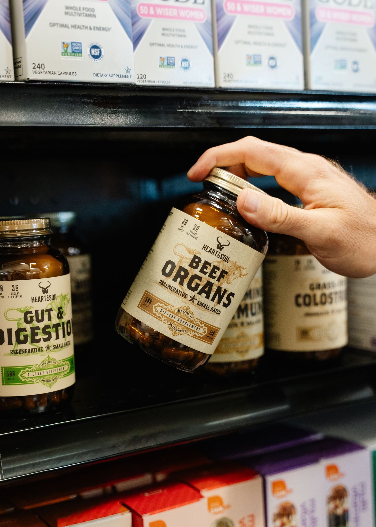 Hand picking a jar of Heartland Beef Organs supplement from a retail shelf stocked with various health supplements.