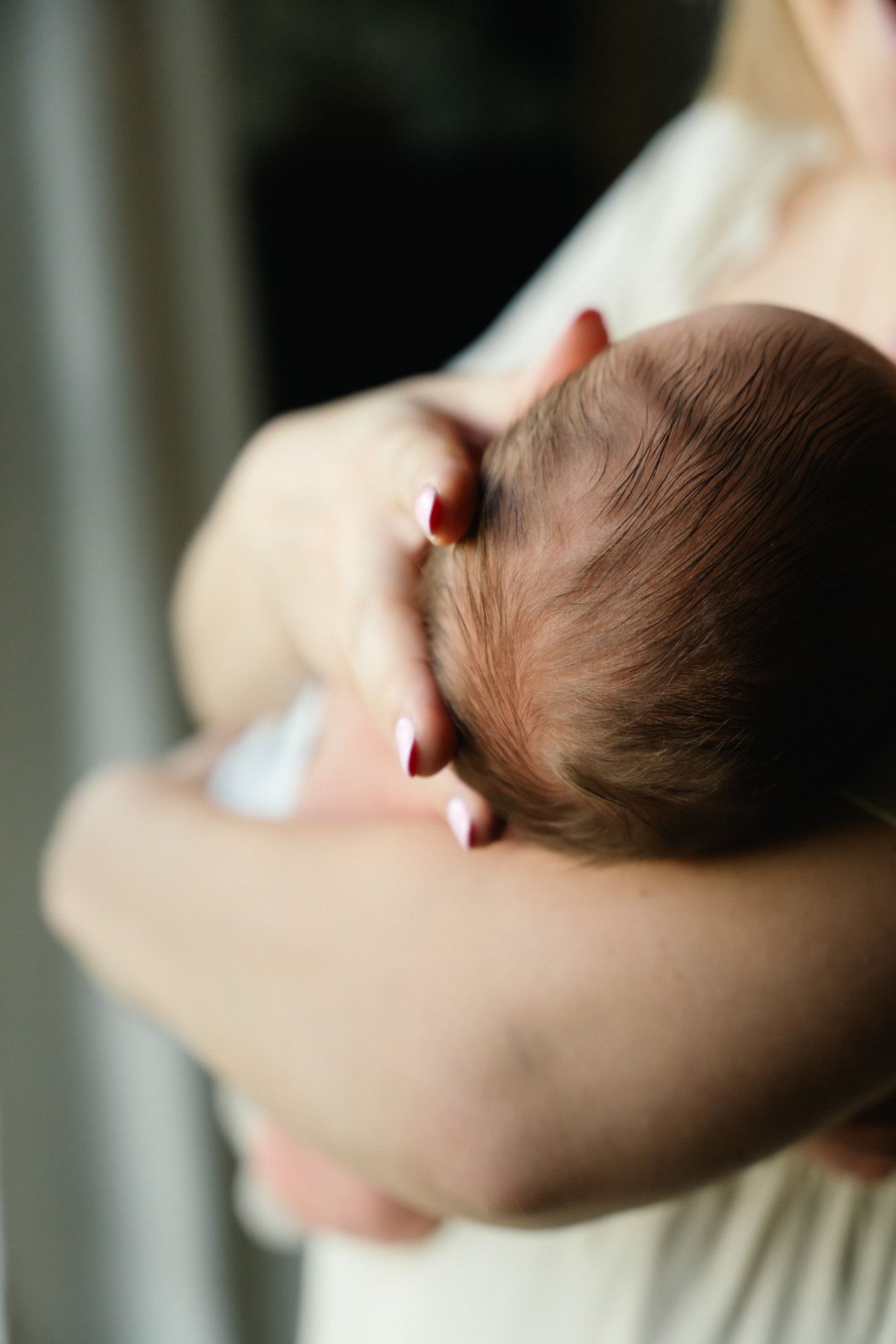 Close-up of a newborn baby's head with dark hair cradled in mother's hands with red nails.