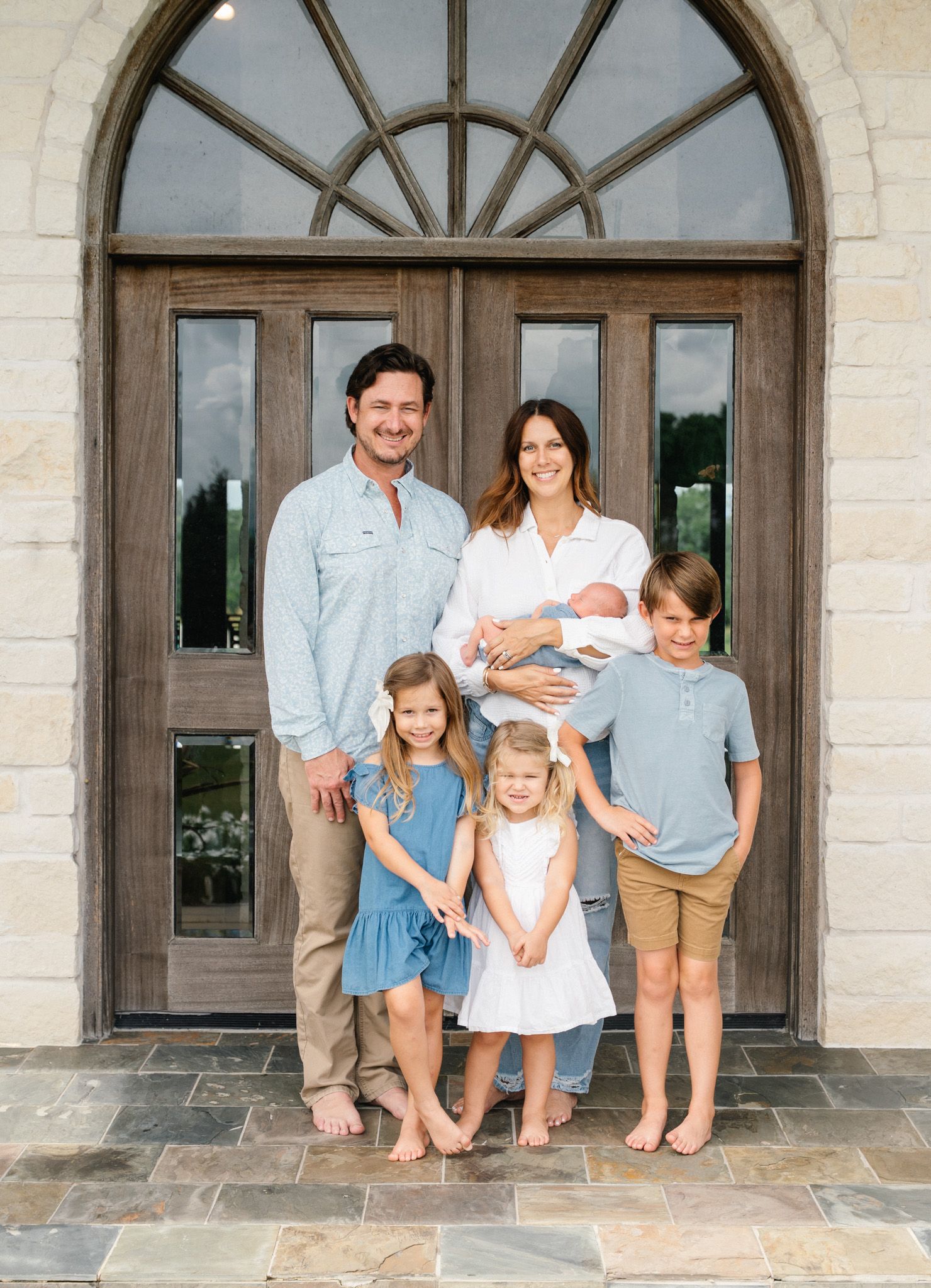Family of six posing with newborn baby in front of elegant stone and wood double doors.
