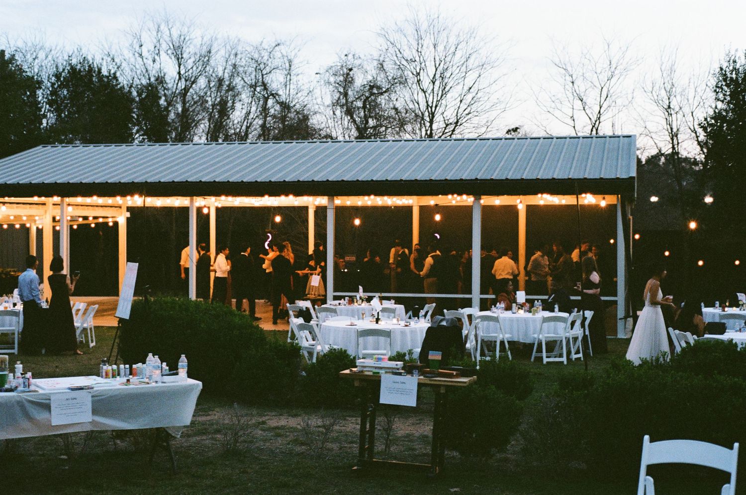 Outdoor wedding reception venue with string lights, white tables, and guests mingling at dusk.