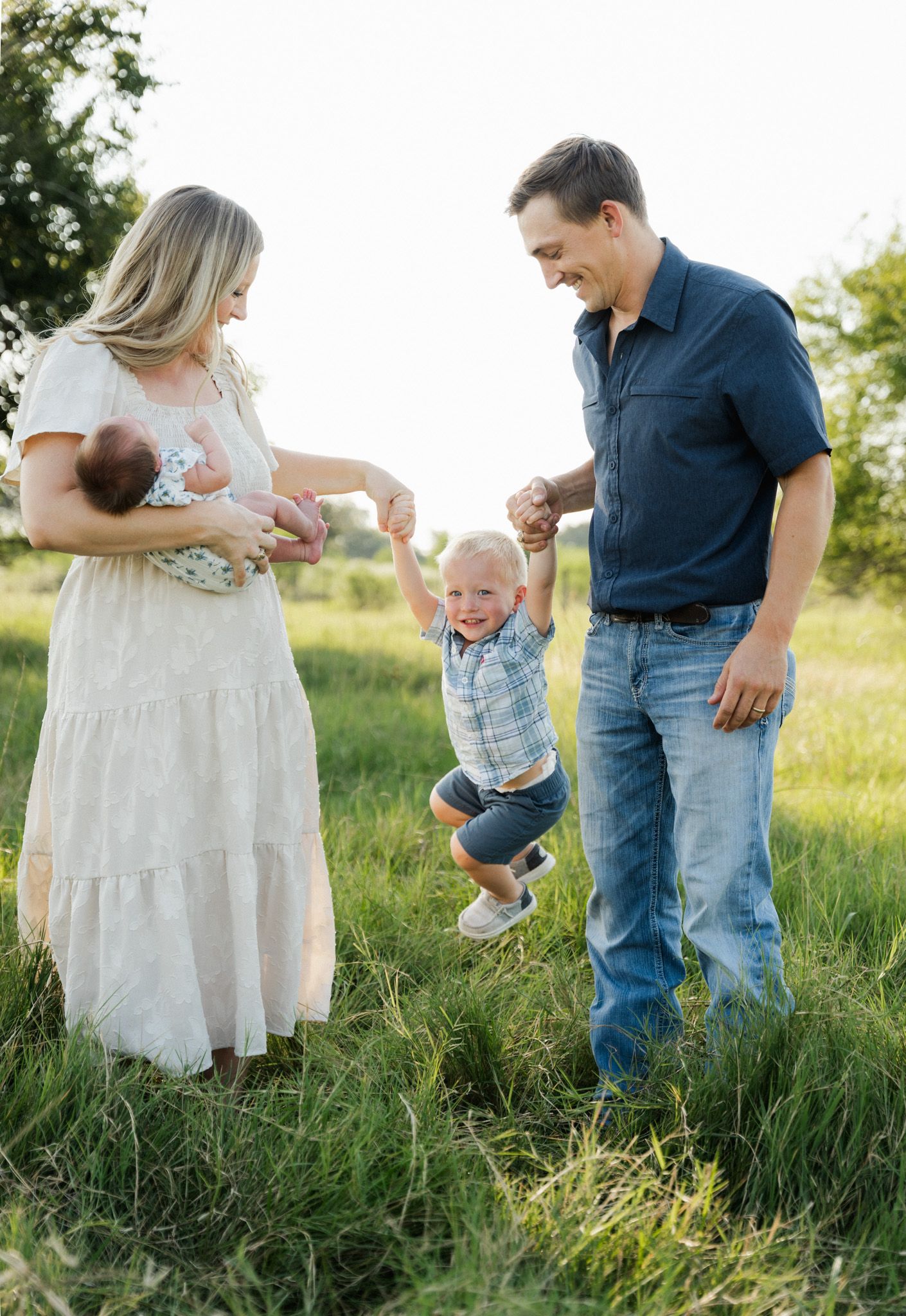A joyful family of four in a sunny green field, parents swinging toddler while mom holds newborn baby.