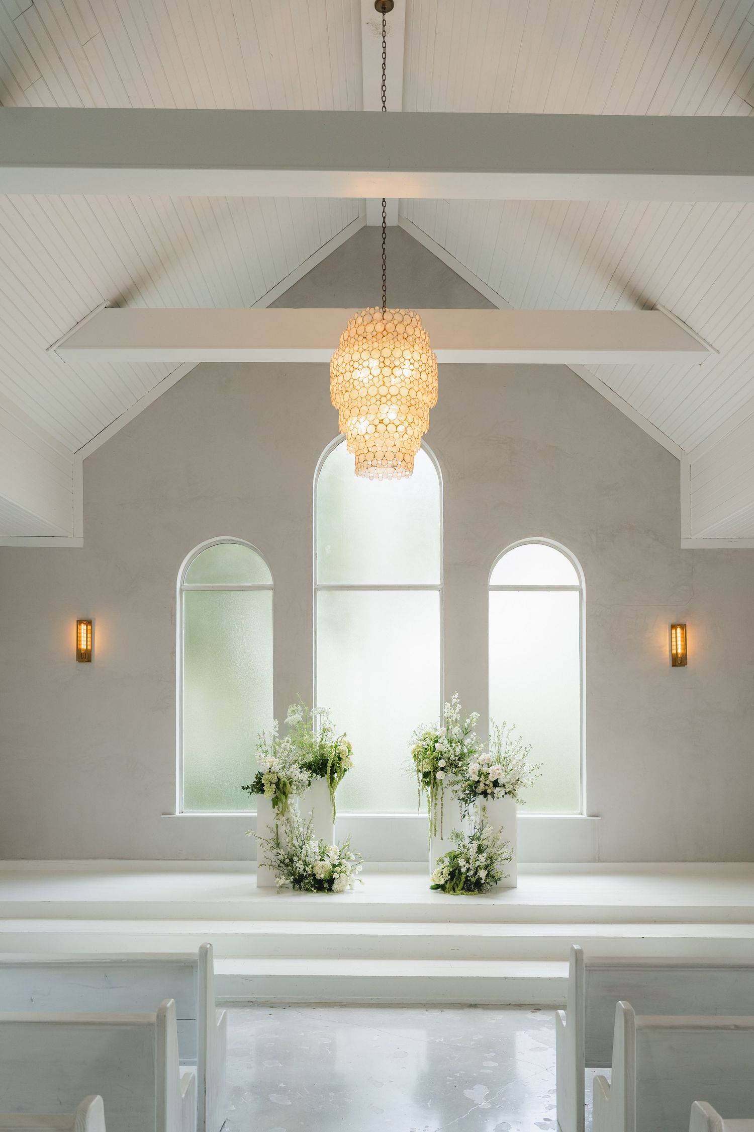 Elegant white chapel interior with vaulted ceiling, crystal chandelier, arched windows, and floral arrangements on altar.
