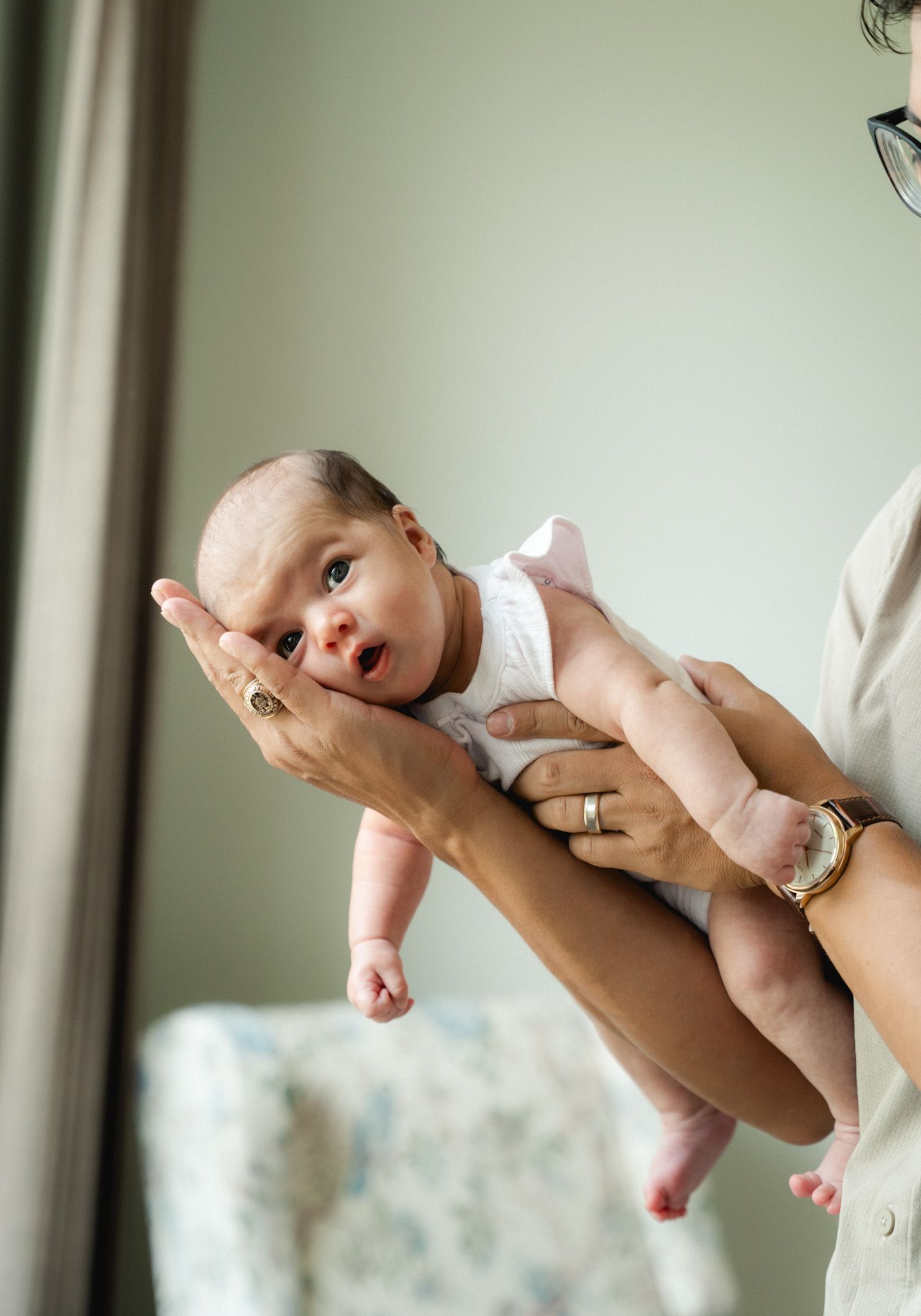 Adult hands gently holding a wide-eyed newborn baby in a white onesie, lifted up near a bright window.