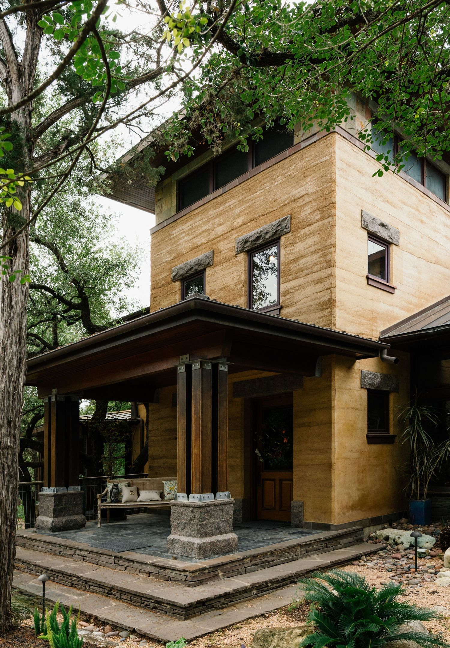 Two-story rammed earth house with covered porch, stone columns, dark wood beams, surrounded by mature trees.