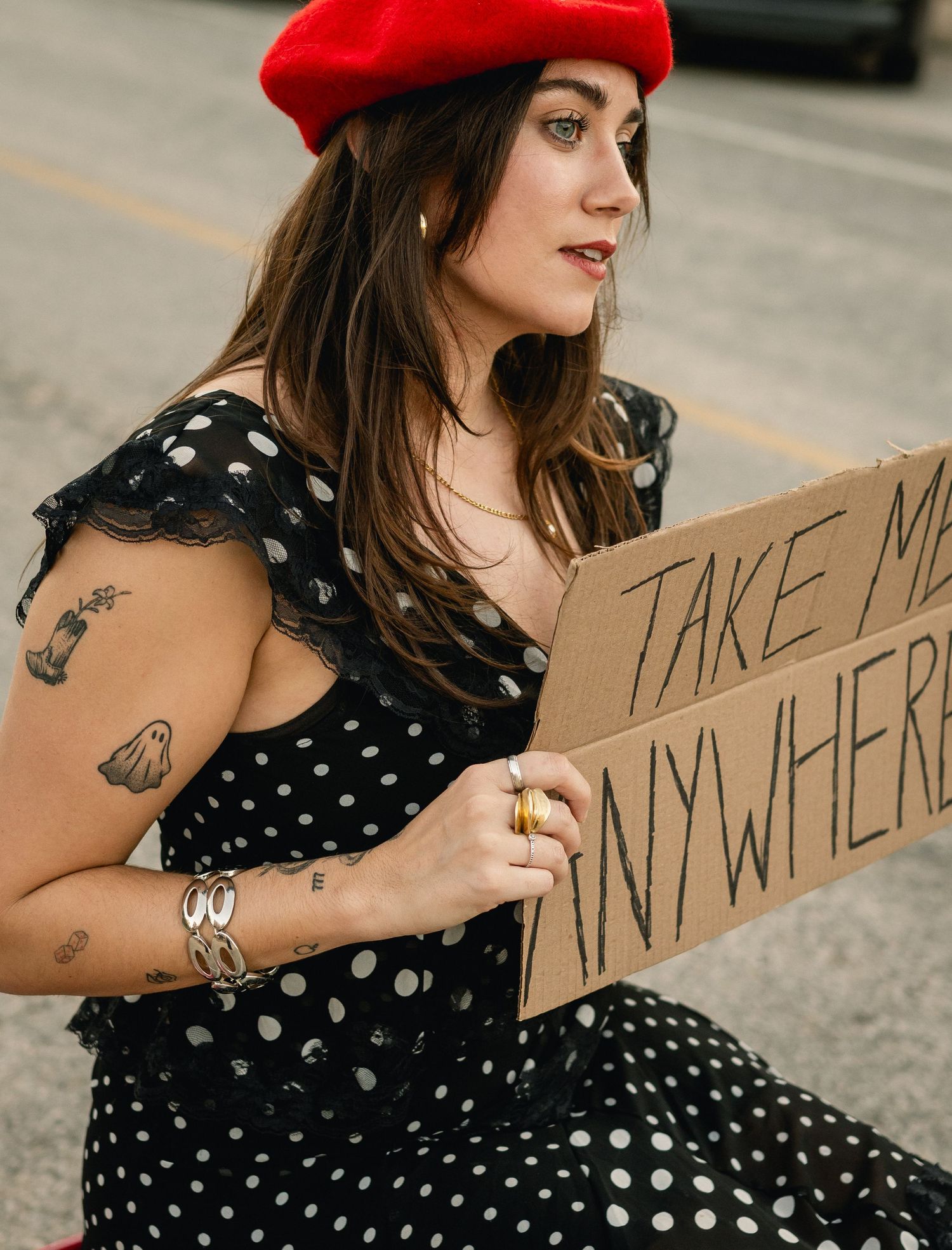 Woman in polka dot dress and red beret holding cardboard sign reading Take Me Anywhere.