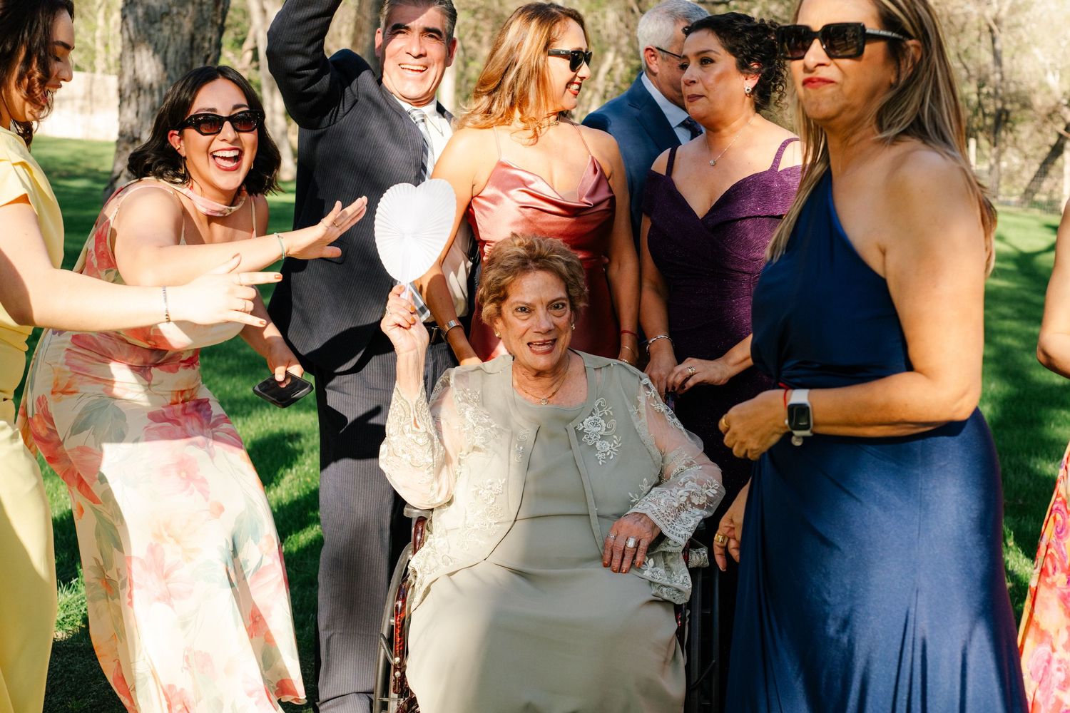 Group of elegantly dressed wedding guests surrounding a woman in a wheelchair, laughing and celebrating outdoors.