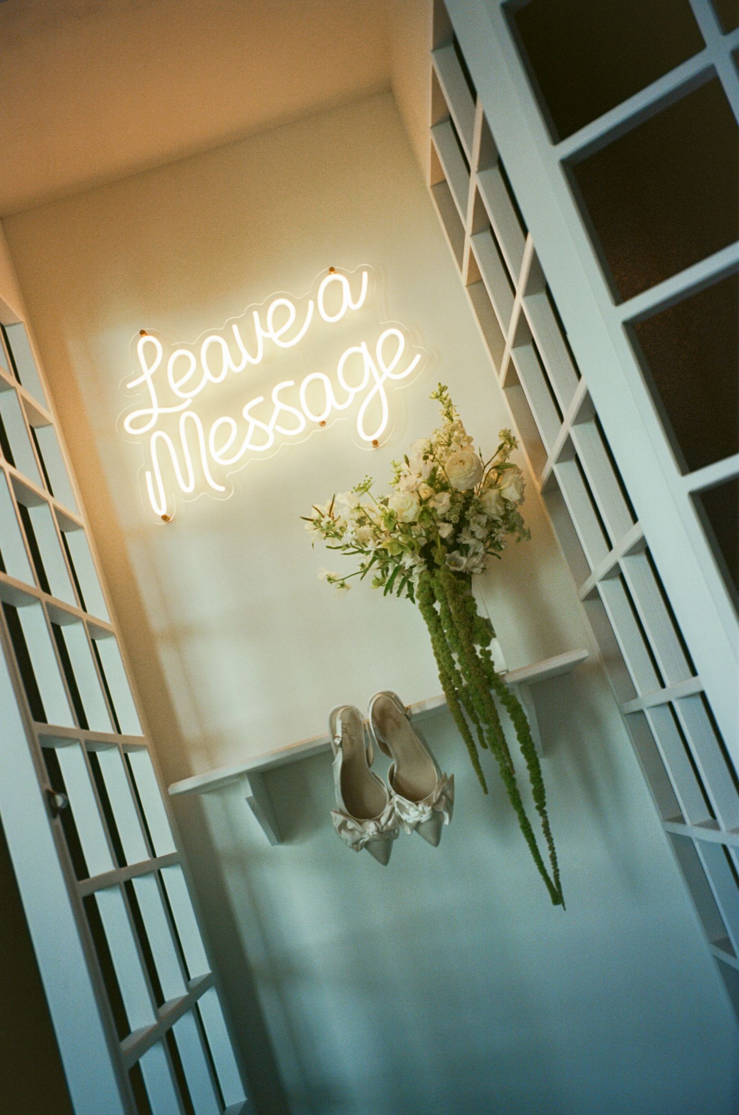 Bridal heels and white floral bouquet displayed on shelf beneath glowing neon Leave a Message sign.