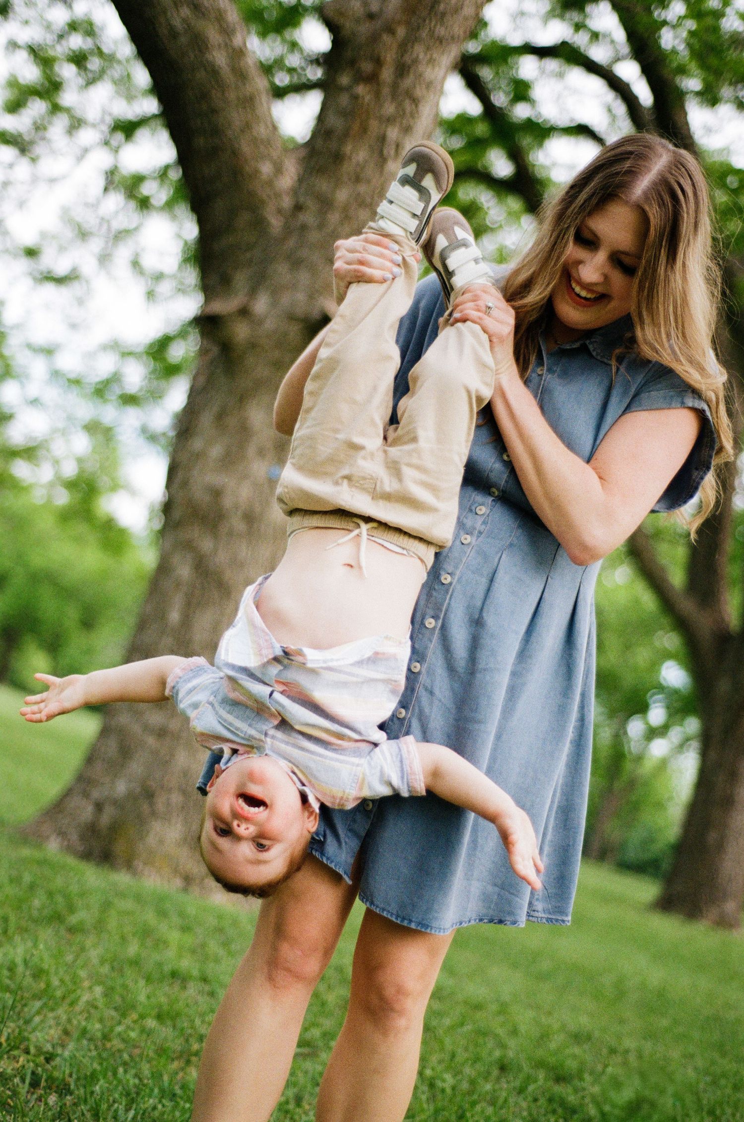 Smiling mother playfully holding her laughing toddler upside down outdoors near large trees.